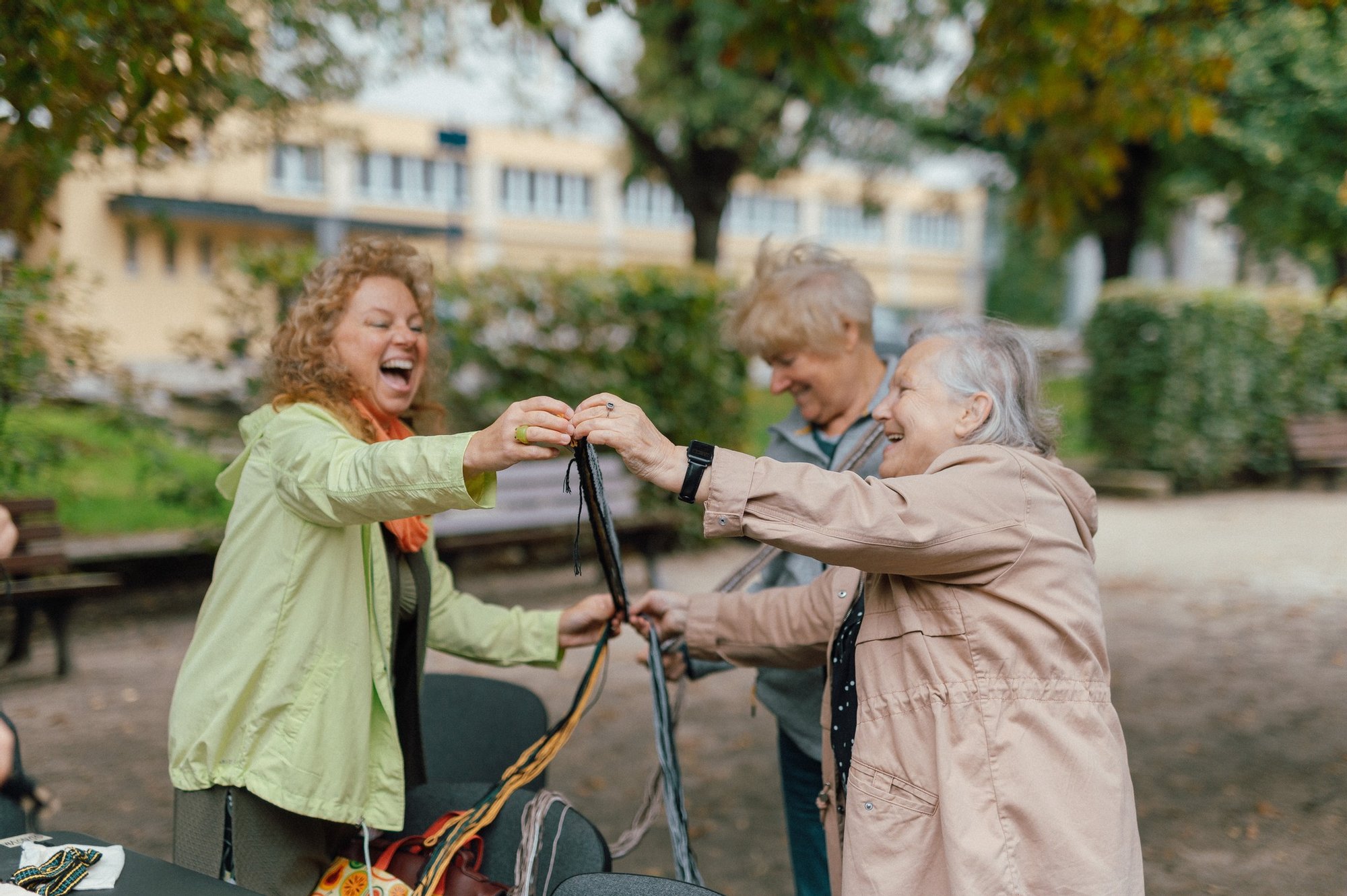Trzy roześmiane osoby - dwie seniorki i kobieta w średnim wieku stoją w parku pod drzewami. Odmierzają długość włóczki do zrobienia paska. Trwają warsztaty rzemiosła.