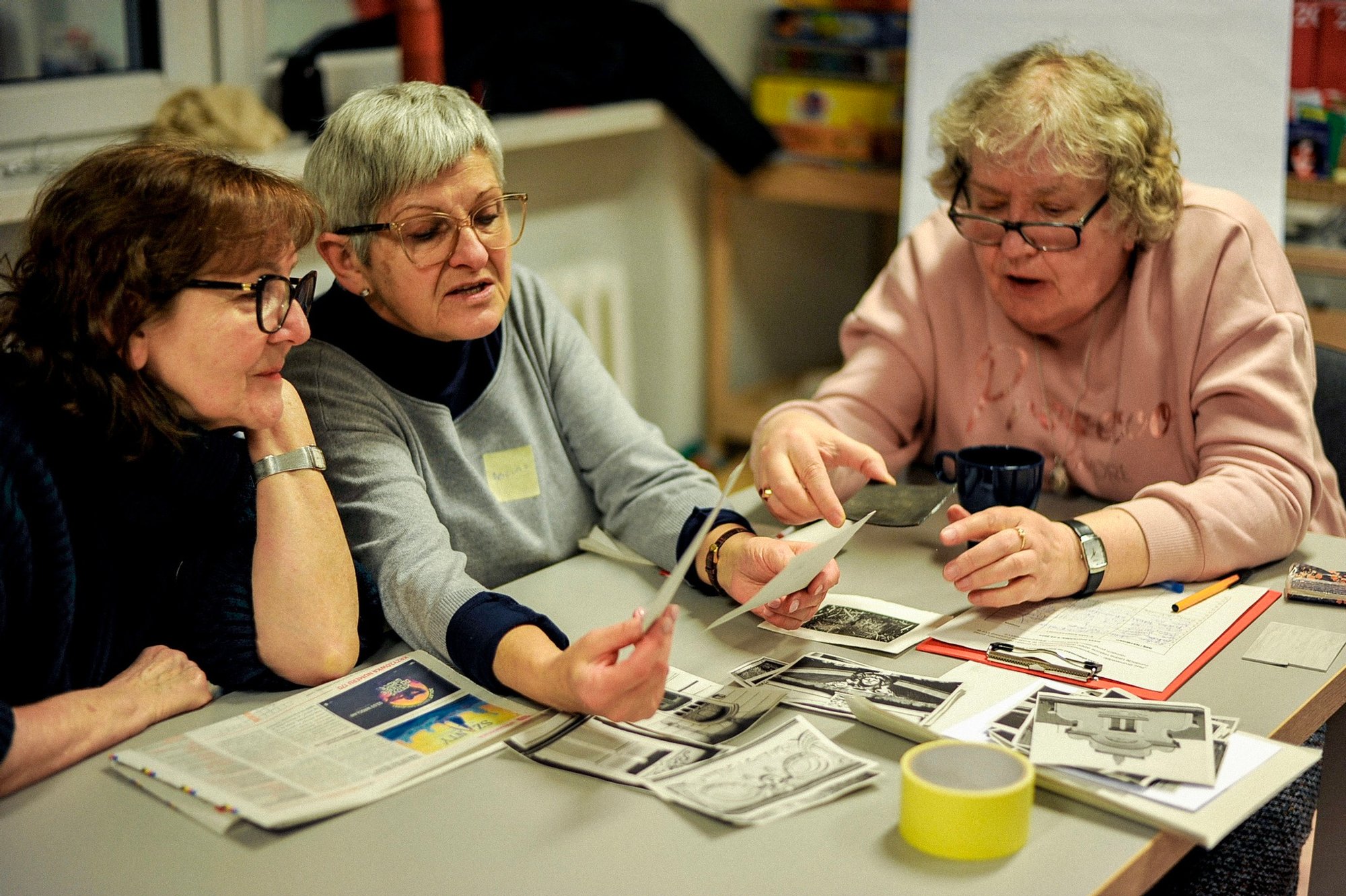 Kolorowa fotografia przedstawiająca trzy seniorki w jasnym pomieszczeniu. Kobiety przeglądają leżące przed nimi na stole archiwalne fotografie.