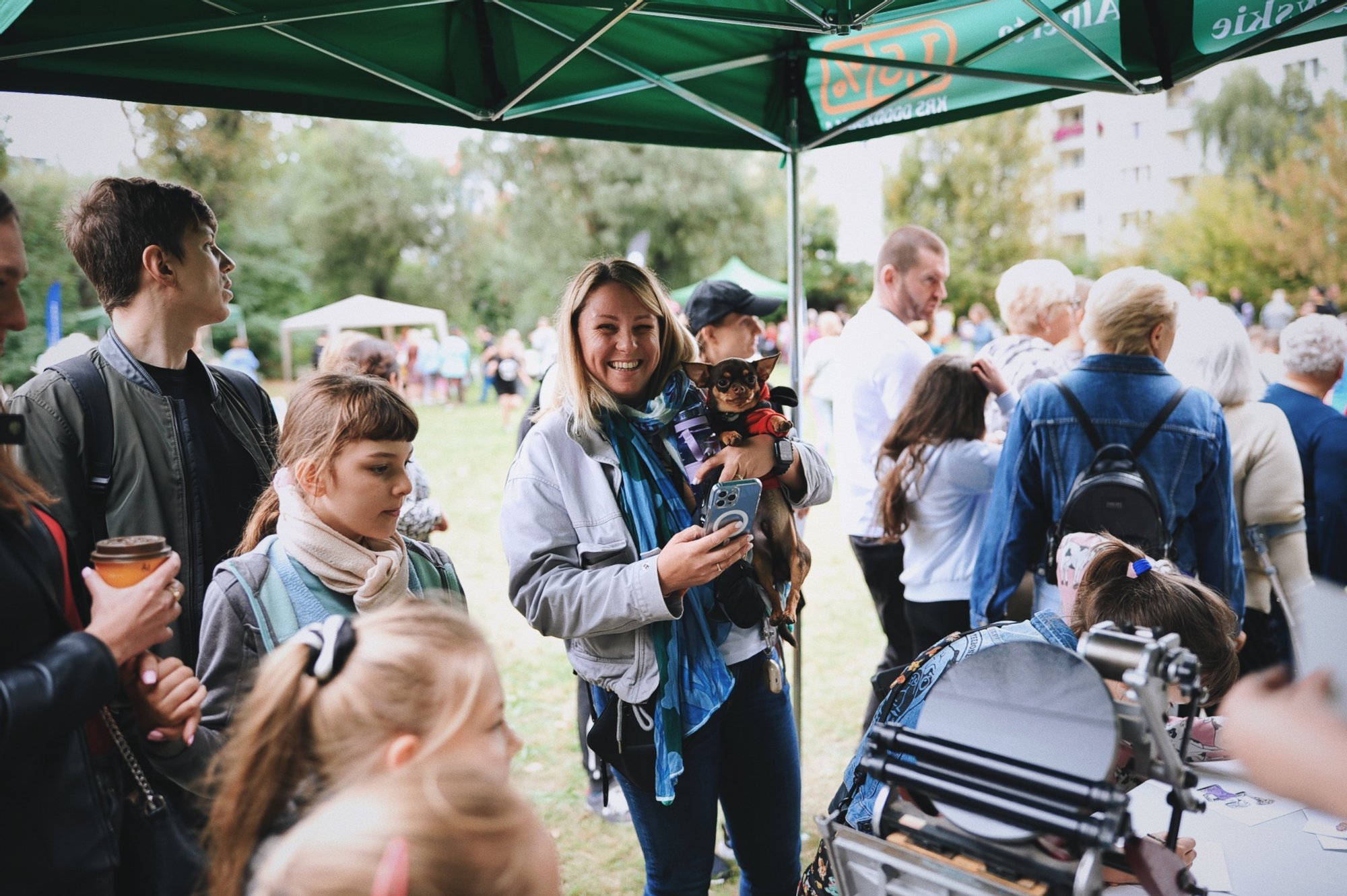 Фото: Анна Паздей Kolorowa fotografia wykonana podczas osiedlowego pikniku. Pod zielonym namiotem rozstawionym na trawie zebrała się grupa osób w różnym wieku. Na środku zdjęcia młoda roześmiana kobieta trzymająca na rękach małego pieska.