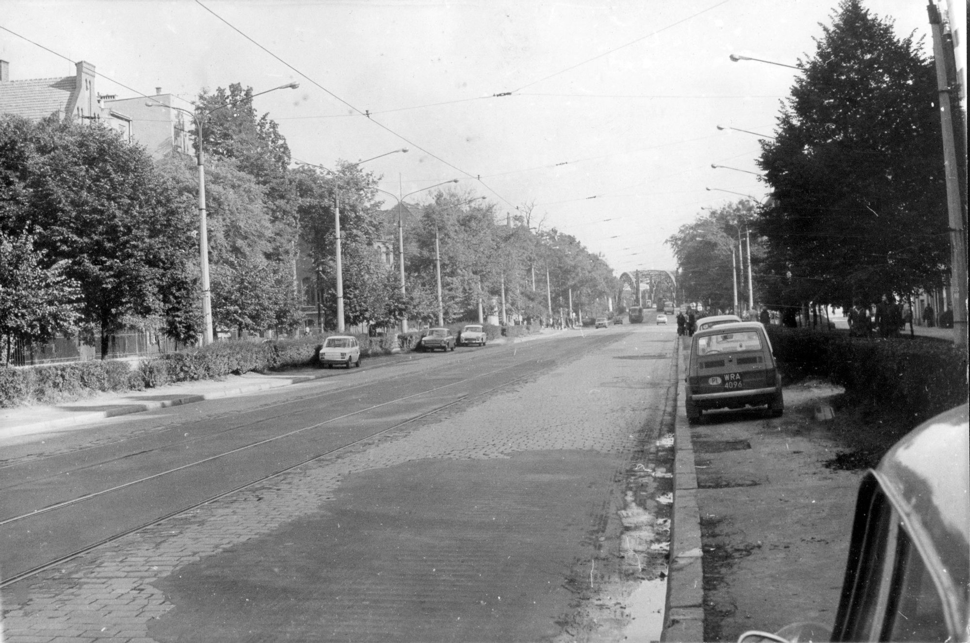 Archiwalne, czarno-białe zdjęcie. Widok na ulicę Curie-Skłodowskiej za skrzyżowaniem z Norwida. Ulica jest pusta, po obu stronach stoi zaparkowanych kilka Fiatów 126p.