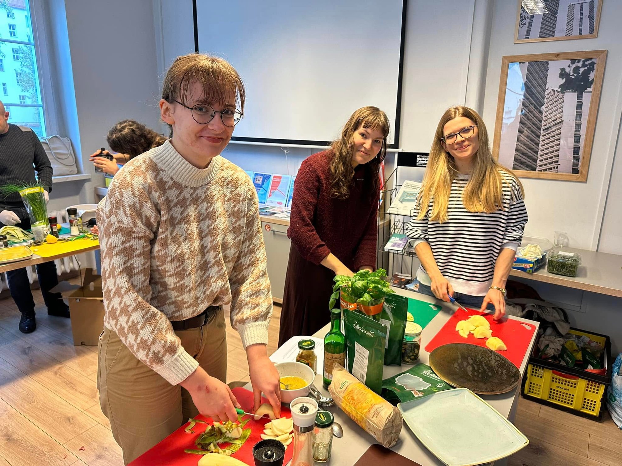 Kolorowa fotografia: trzy młode kobiety przygotowują potrawę podczas warsztatów kulinarnych.