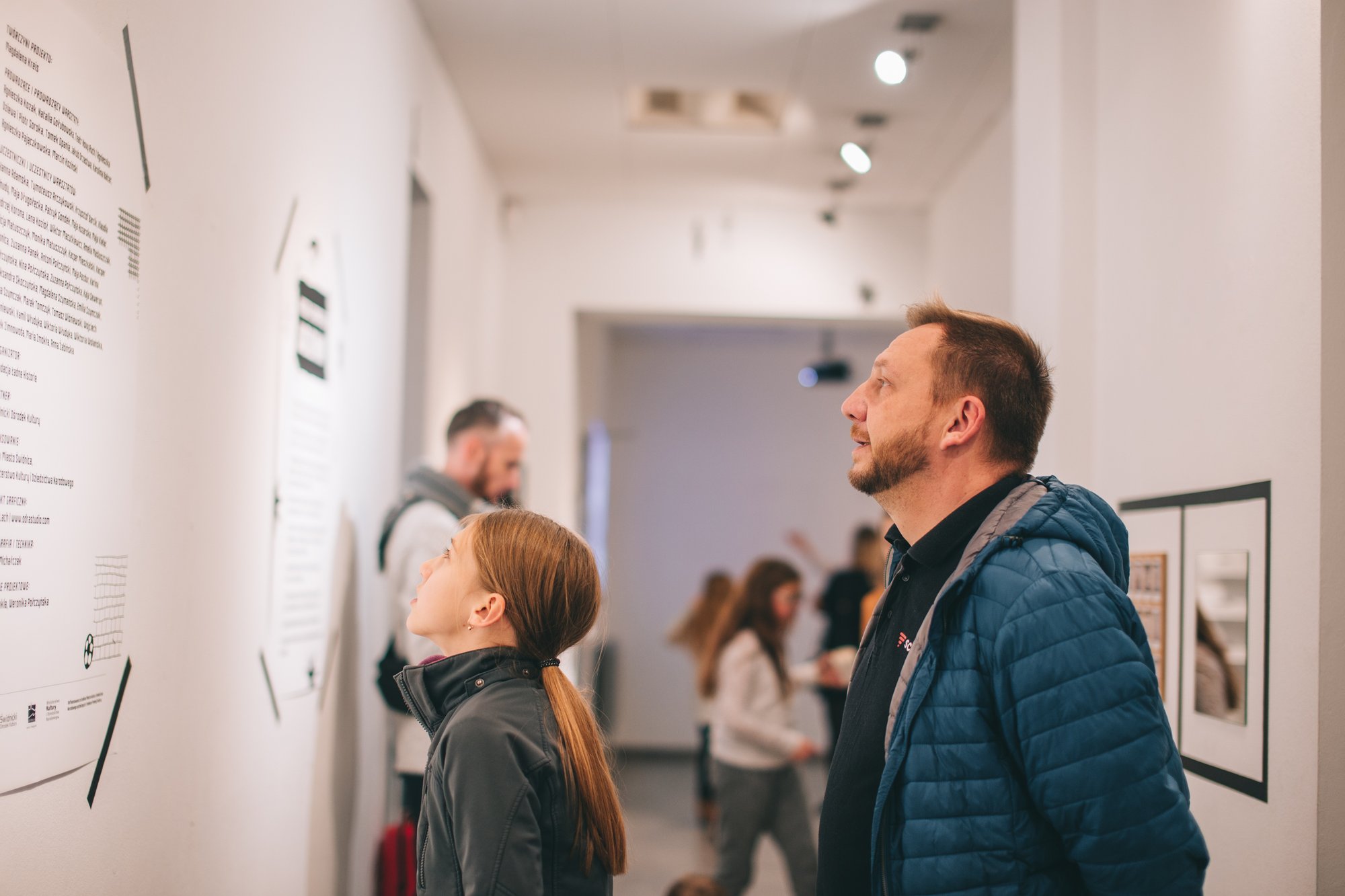 Na ścianach białego pomieśczenia umieszczone elementy wystawy. Na jeden z nim zapatrzona jest młoda dziewczyna w ciemnej kurtce, z włosami związanymi w kucyk. Za nią stoi starszy mężczyzna, również z uwagą czyta treść plakatu.