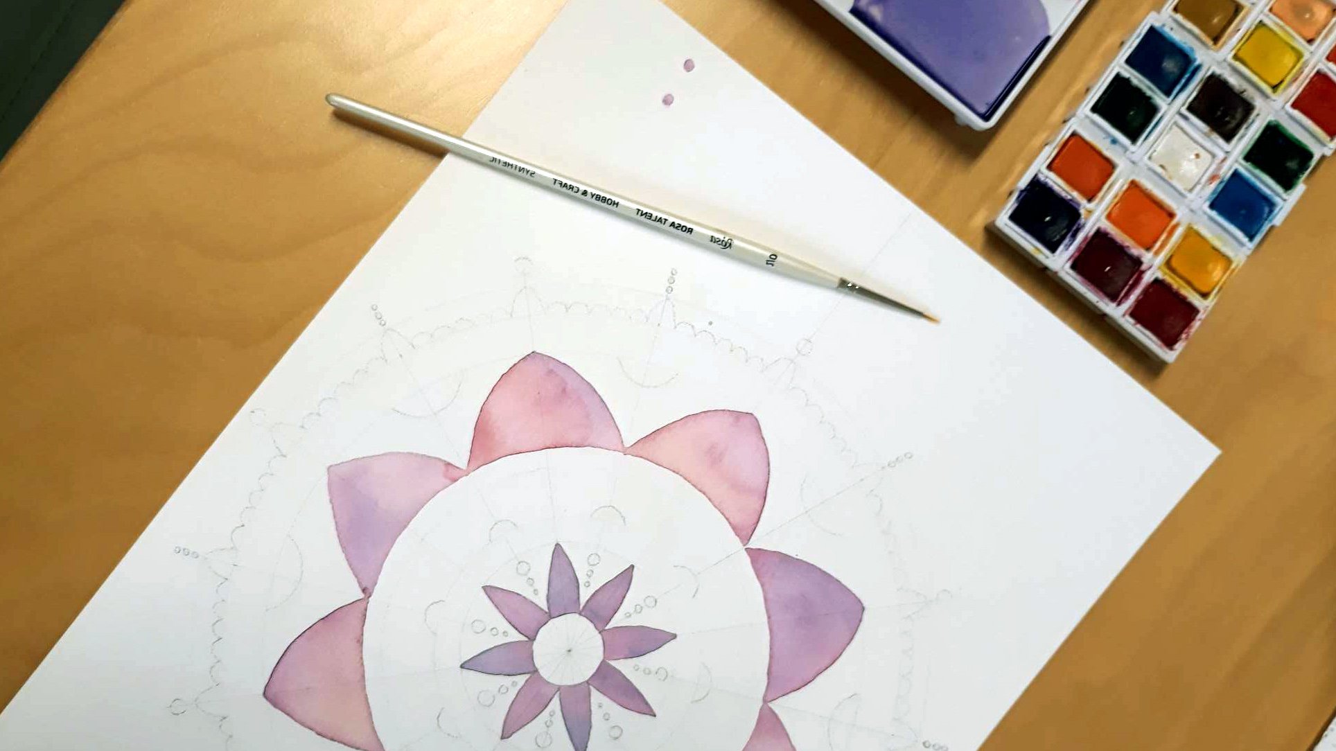 fot. Alona Bondarenko Widok z góry na leżący na stole rozpoczęty rysunek mandali. Obok pędzelek i pudełko farb,