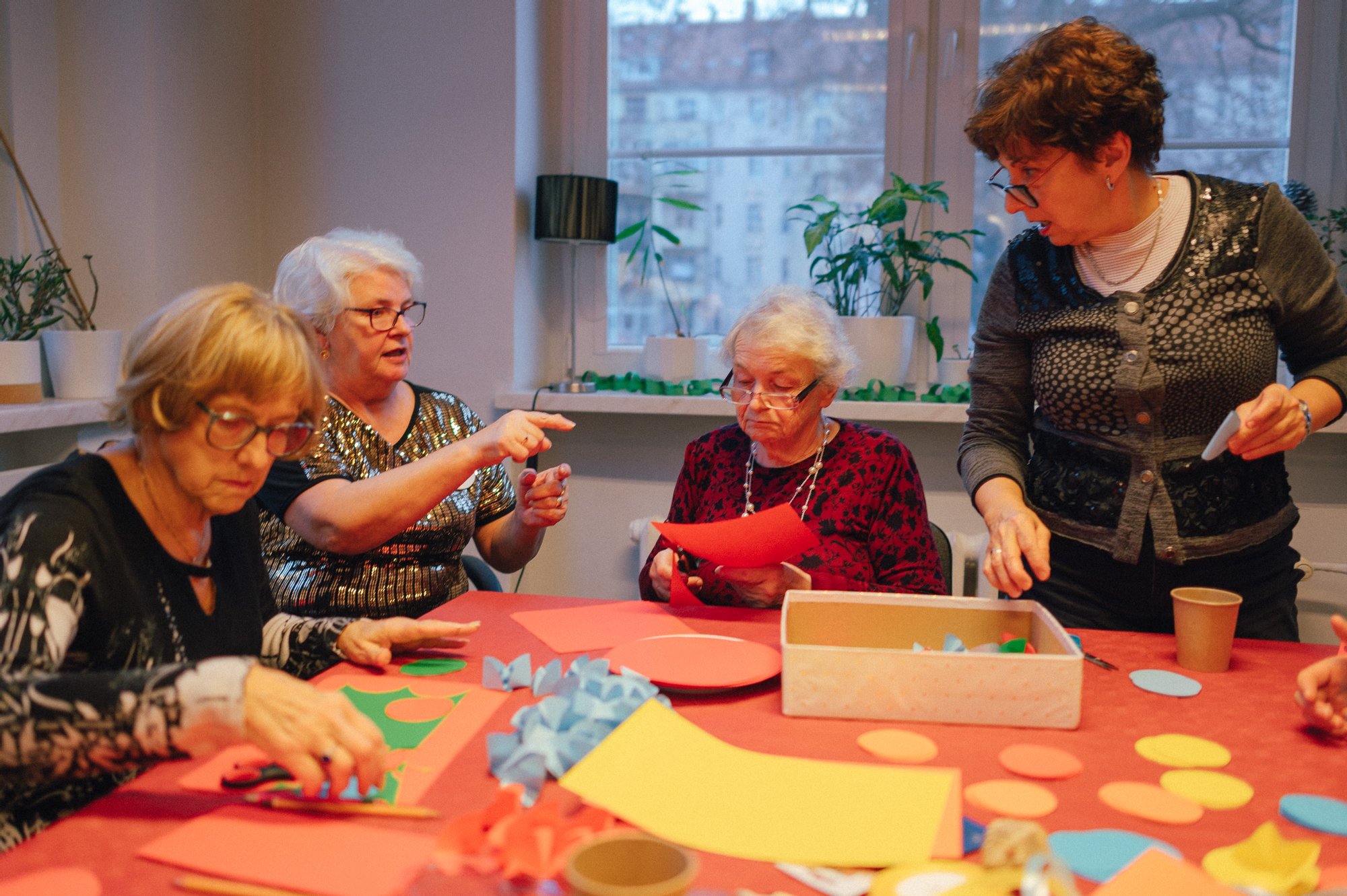 Kolorowa, pozioma fotografia: w jasnym pomieszczeniu przy stole siedzą cztery seniorki, przed nimi rozłożone są materiały plastyczne. Kobiety robią ozdoby świąteczne.