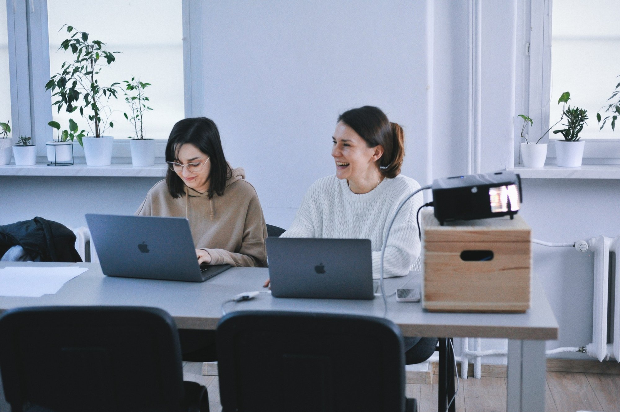 Kolorowa, pozioma fotografia: w jasnym pomieszczeniu przy stole siedzą dwie młode, roześmiane kobiety. Przed nimi na stole leżą otwarte laptopy.