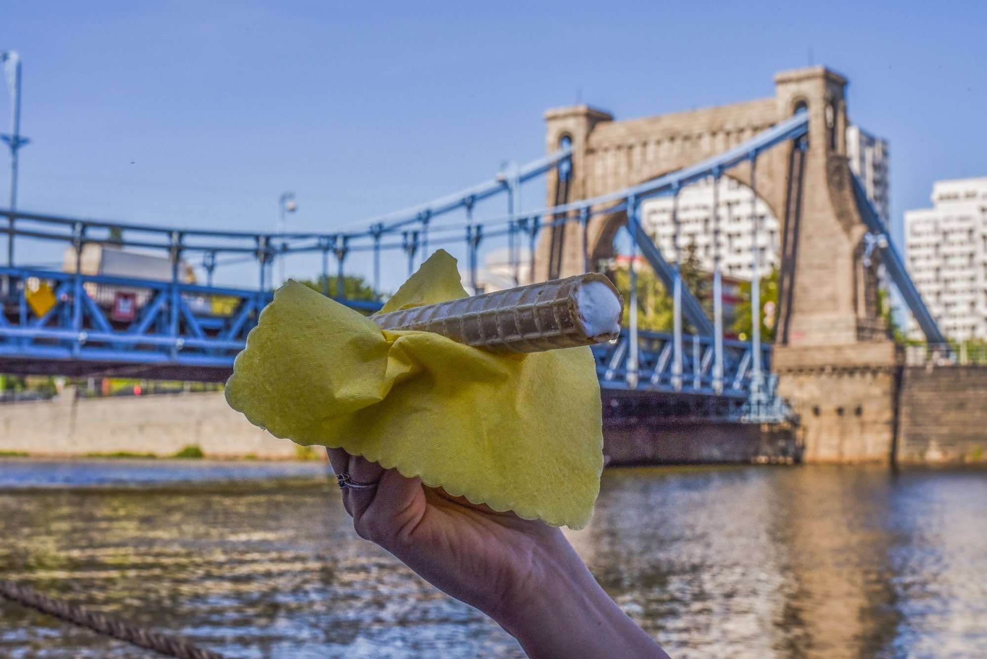 Kolorowa, pozioma fotografia. Zbliżenie na dłoń trzymającą przez żółtą serwetkę rurkę z kremem. W tle Most Grunwaldzki.  