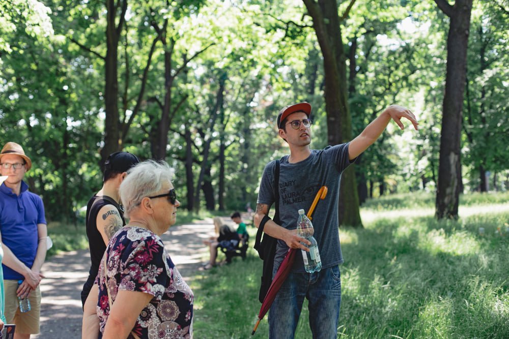 fot. Marta Sobala Przewodnik Maciej Wlazło stoi z grupą ludzi w parku. W jednej ręce trzyma zamknięty parasol i dużą butelkę wody, drugą ręką wskazuje coś poza kadrem. W tle widoczne wysokie drzewa oraz szeroka ścieżka.