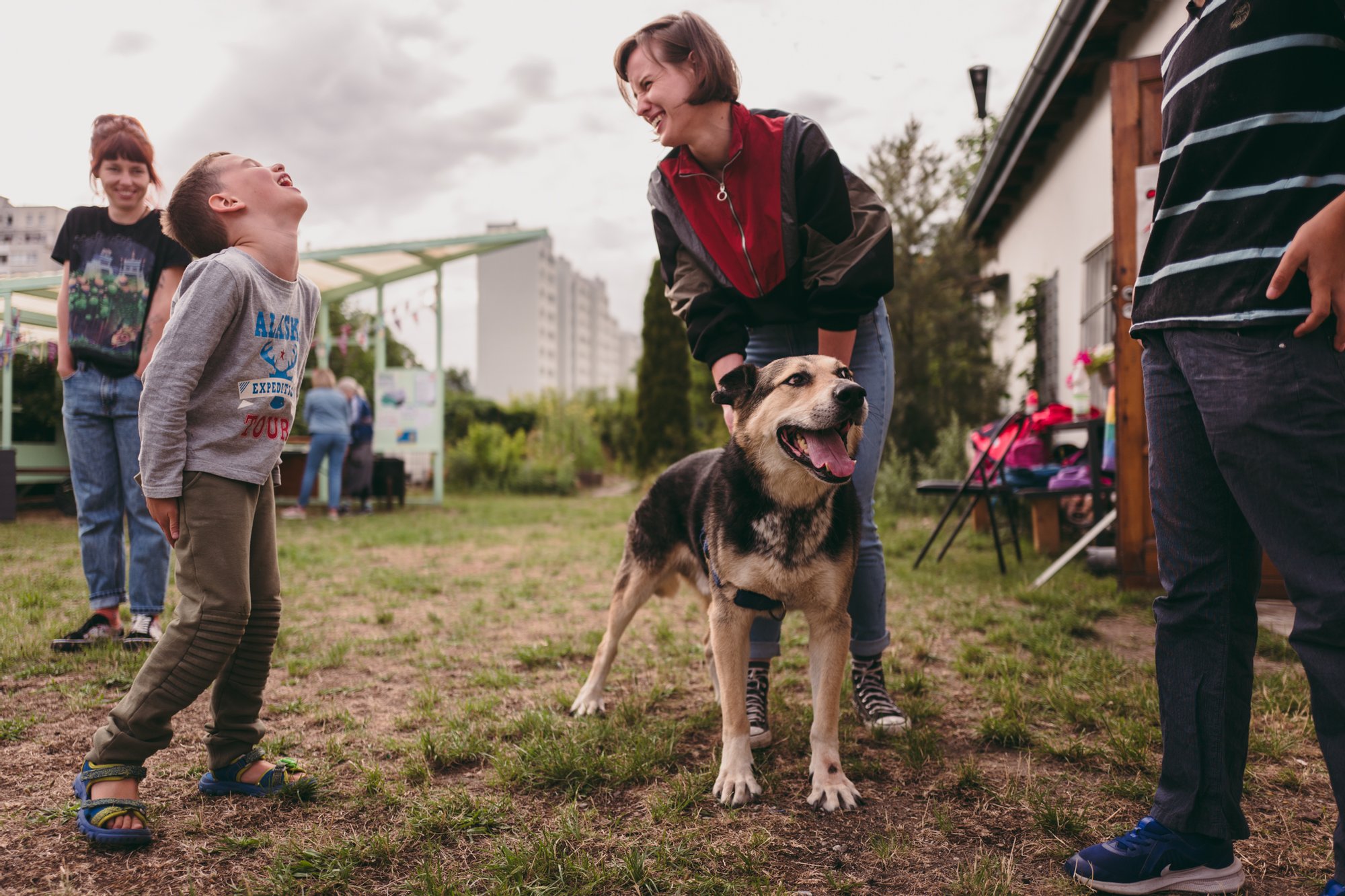 Fot. Wojciech Chrubasik Kolorowa, pozioma fotografia przedstawiająca kobietę, głaszczącą psa oraz dziecko na terenie ogródka działkowego. Oboje śmieją się.
