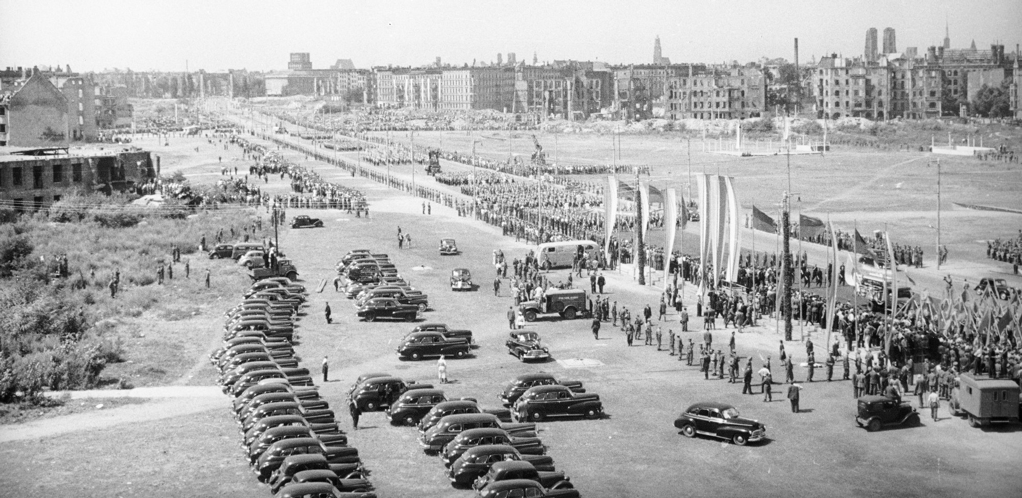 Czarno-biała archiwalna fotografia przedstawia Plac Grunwaldzki w dniu Kongresu Jedności Młodzieży Polskiej. Na zdjęciu widoczne są dziesiątki samochodów równo zaparkowanych oraz tysiące maszerujących osób.