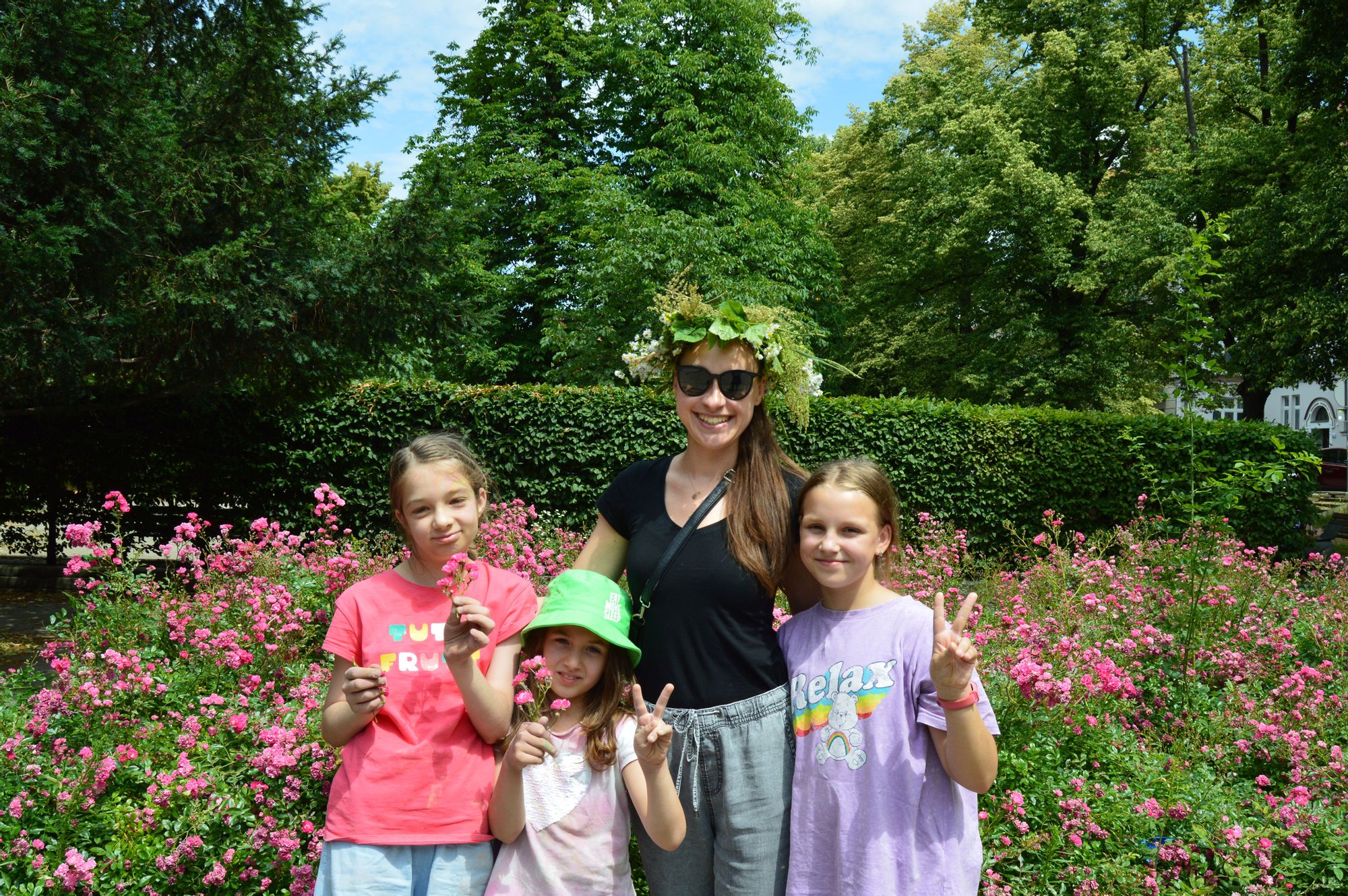 Kolorowa fotografia: młoda kobieta z kolorowym wiankiem na głowie pozuje do zdjęcia razem z trzema młodymi dziewczynkami na tle bujnego, kolorowo kwitnącego krzaka.