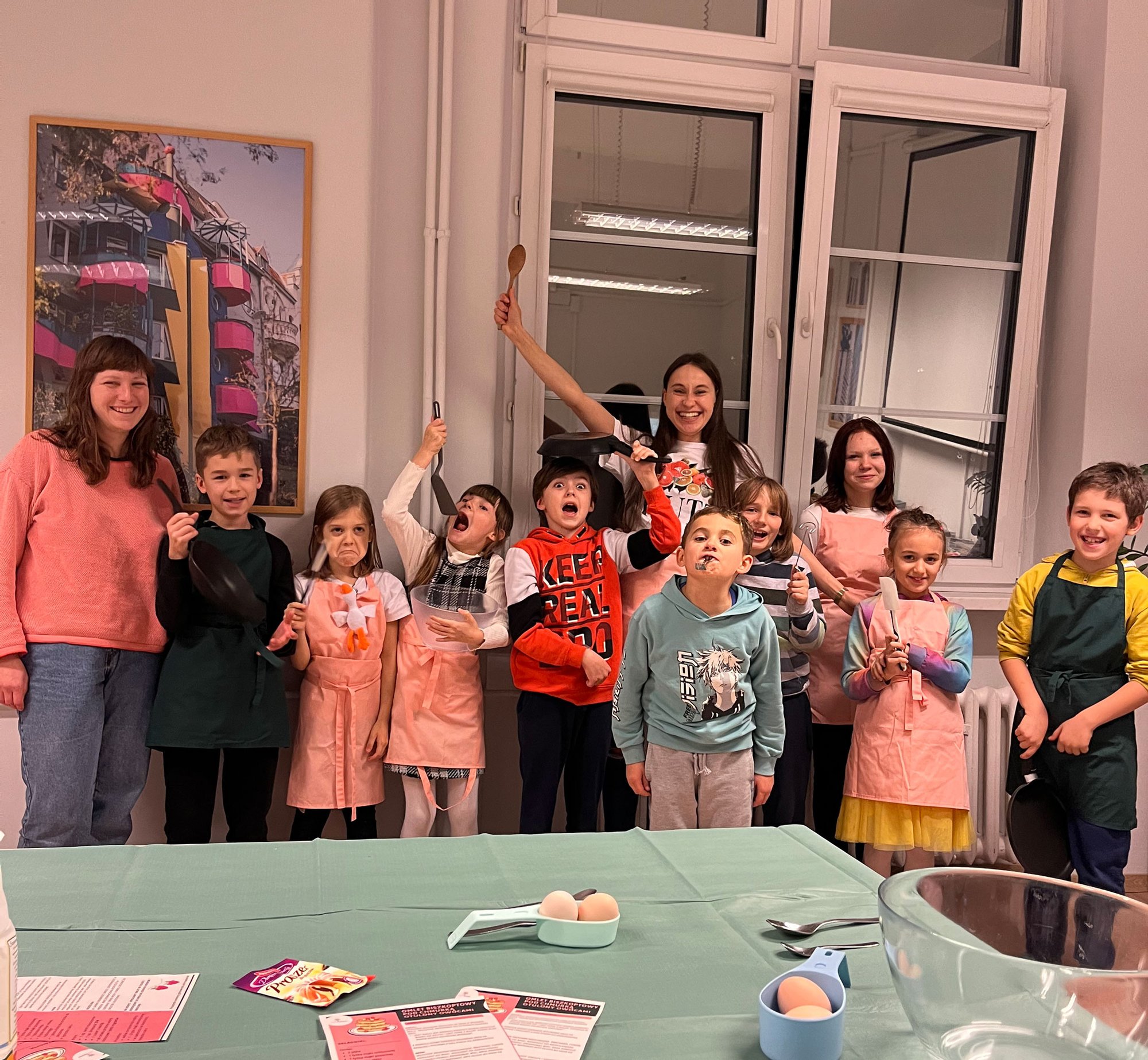 Kolorowa, pozioma fotografia: grupka dzieci w różnym wieku oraz dwie animatorki pozują do zdjęcia w jasnym pomieszczeniu, po zakończonych warsztatach kulinarnych. Dzieciaki machają trzymanymi w rękach łyżkami, chochlami i lizawkami. Na ich twarzach widać radość.
