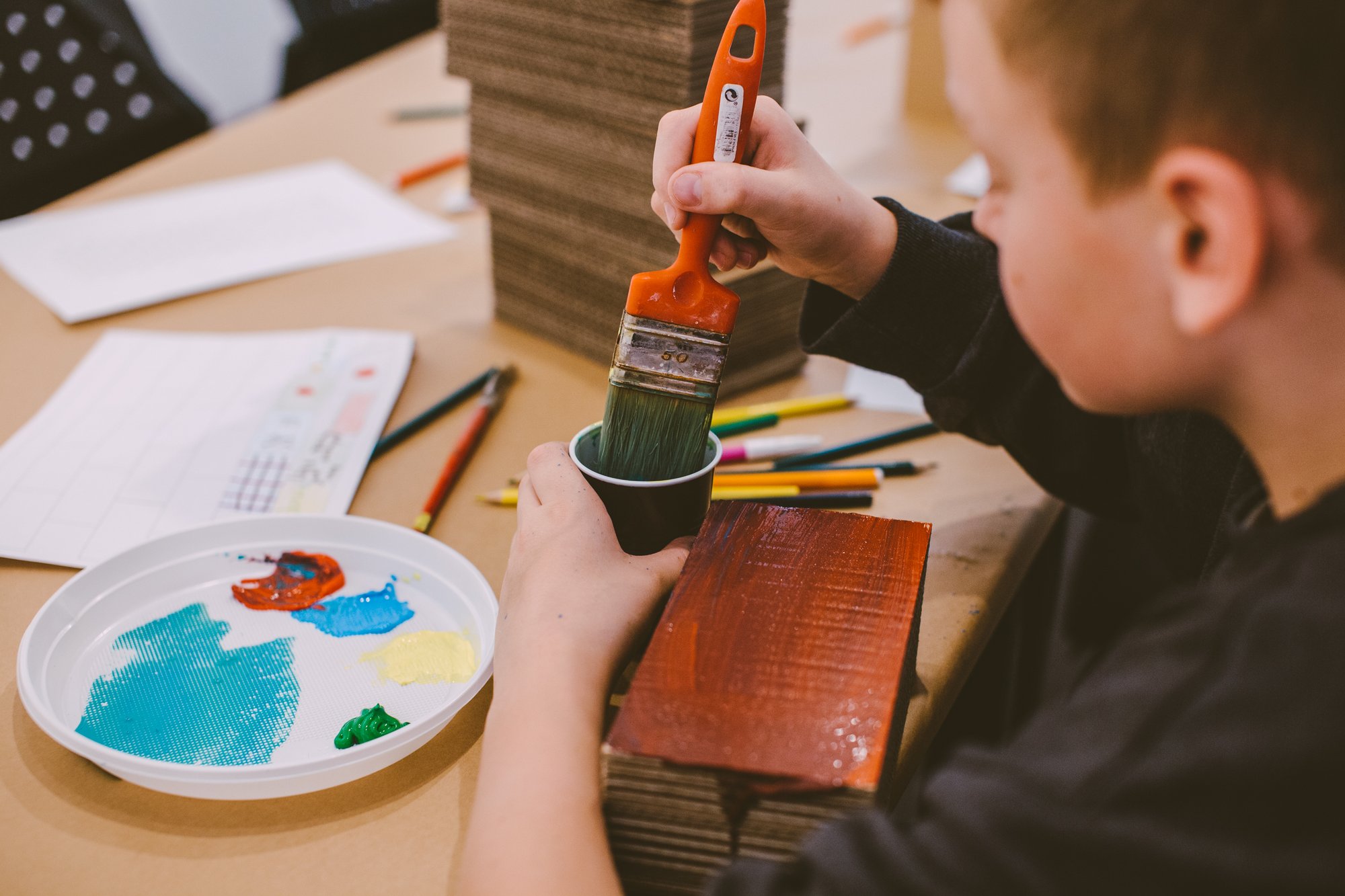 Mały chłopiec siedzi przy stole, na którym rozłożone są kredki, flamastry i kartki papieru. Chłopiec moczy pędzel w kubeczku z wodą. Będzie malował leżące przed nim kawałki drewna.