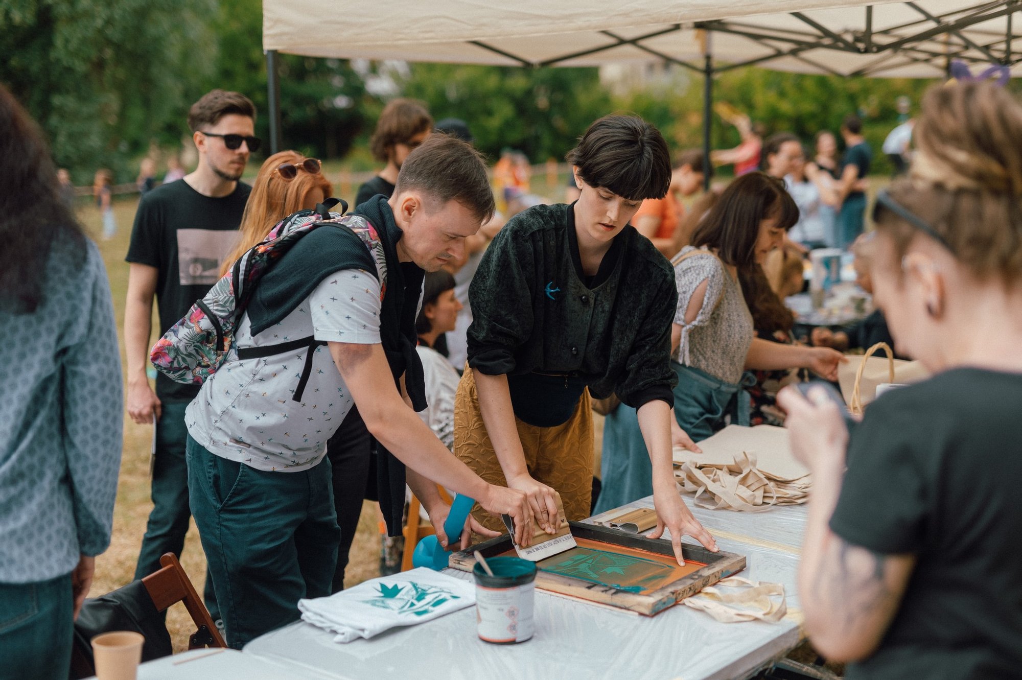 Kolorowa, pozioma fotografia przedstawia ludzi zebranych wokół stołu warsztatowego podczas sąsiedzkiego pikniku. Instruktorka pokazuje jednemu z mieszkańców jak nadrukować wzór metodą sitodruku na lnianej torbie.
