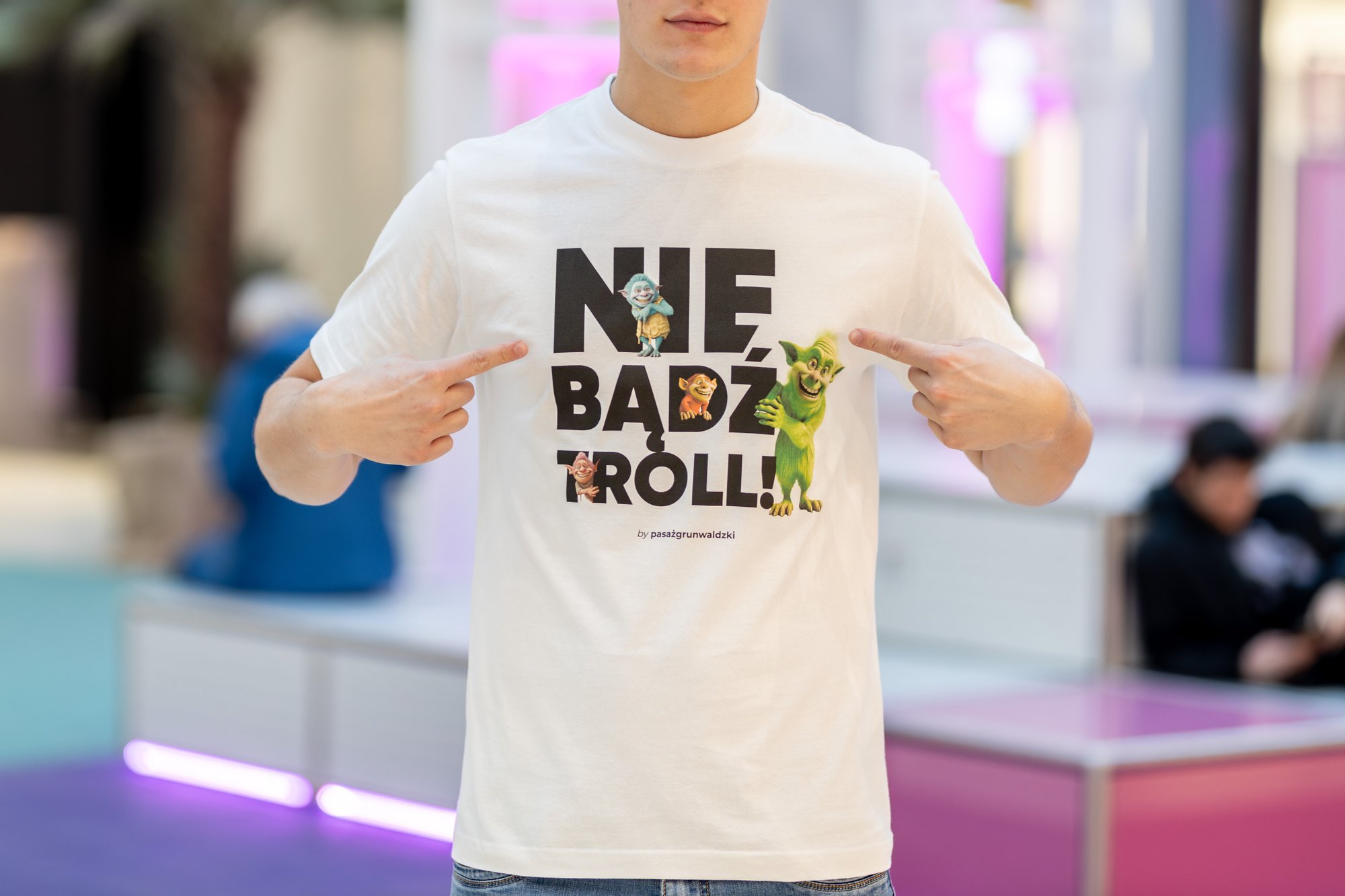Kolorowa, pozioma fotografia: Zbliżenie na białą koszulkę młodego chłopaka. Na koszulce czarny napis "nie bądź troll!" oraz gnomów z filmu animowanego. Chłopak wskazuje palcami na napis.