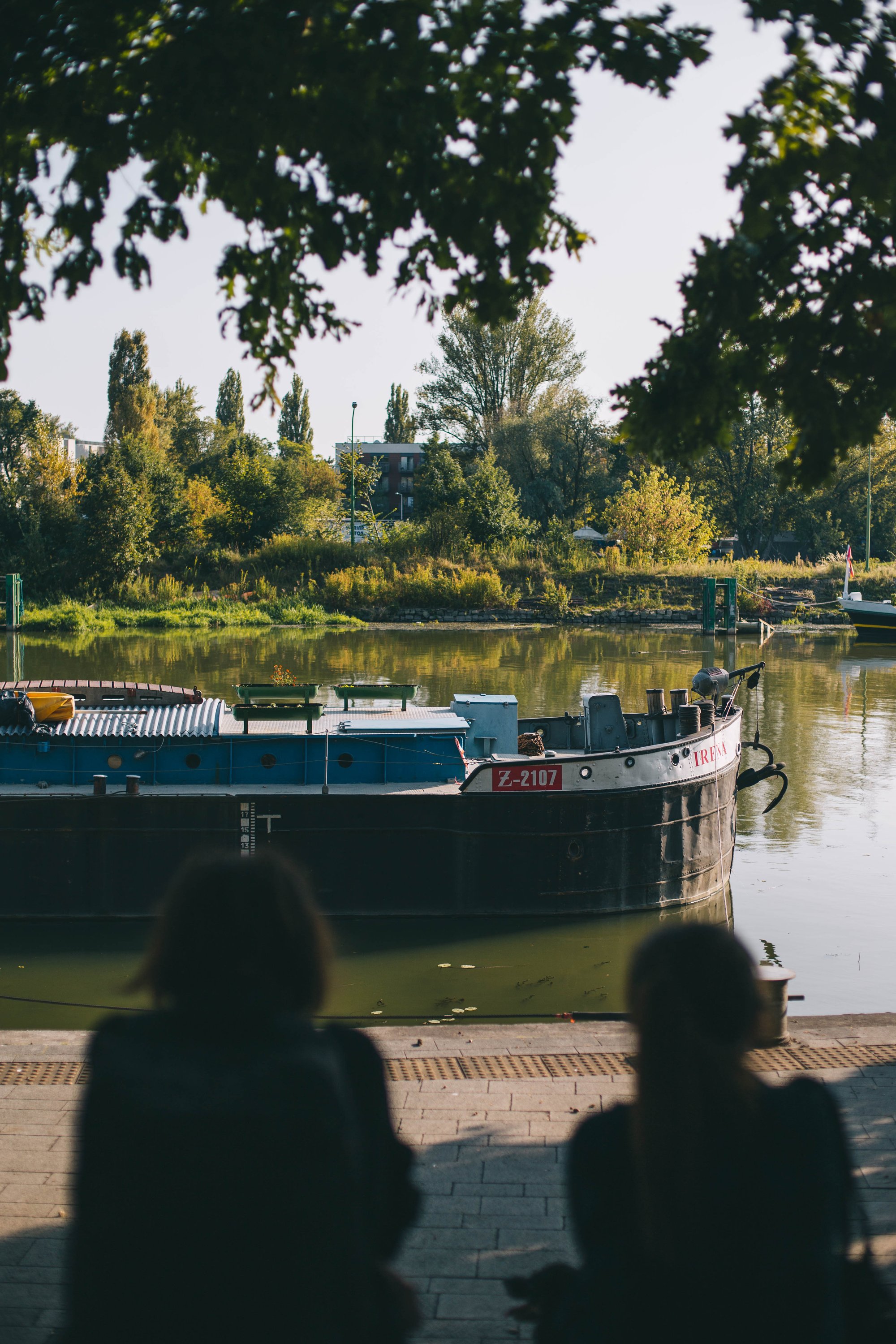 Na pierwszym planie nieostre sylwetki dwóch kobiet siedzących na wybrzeżu Wyspiańskiego. Siedzą patrząc się na barkę przycumowaną na Odrze.