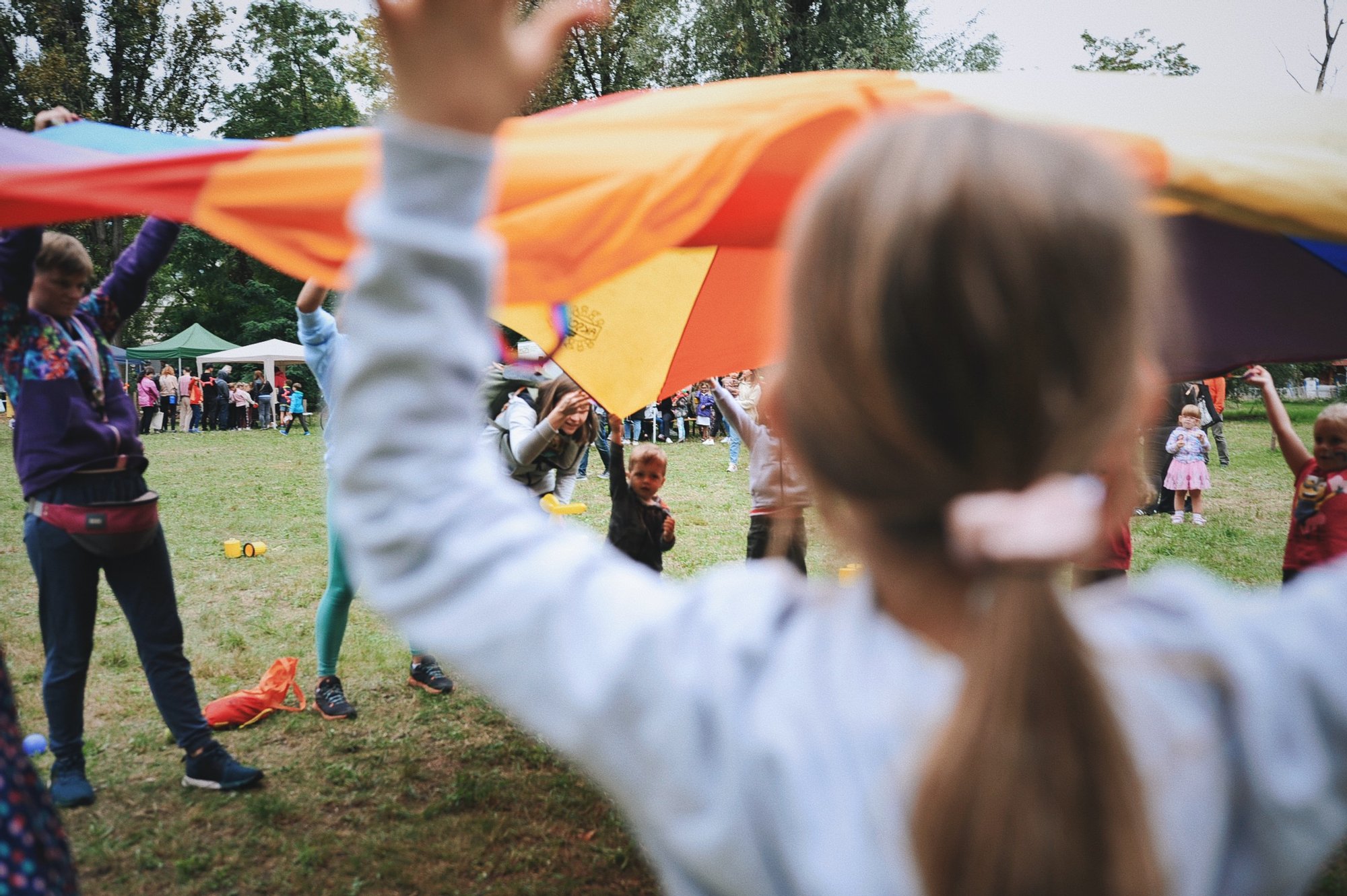zdjęcie: Anna Pazdej Kolorowa fotografia wykonana podczas osiedlowego pikniku. Grupa dzieci wraz z animatorkami bawią się kolorową chustą animacyjną.