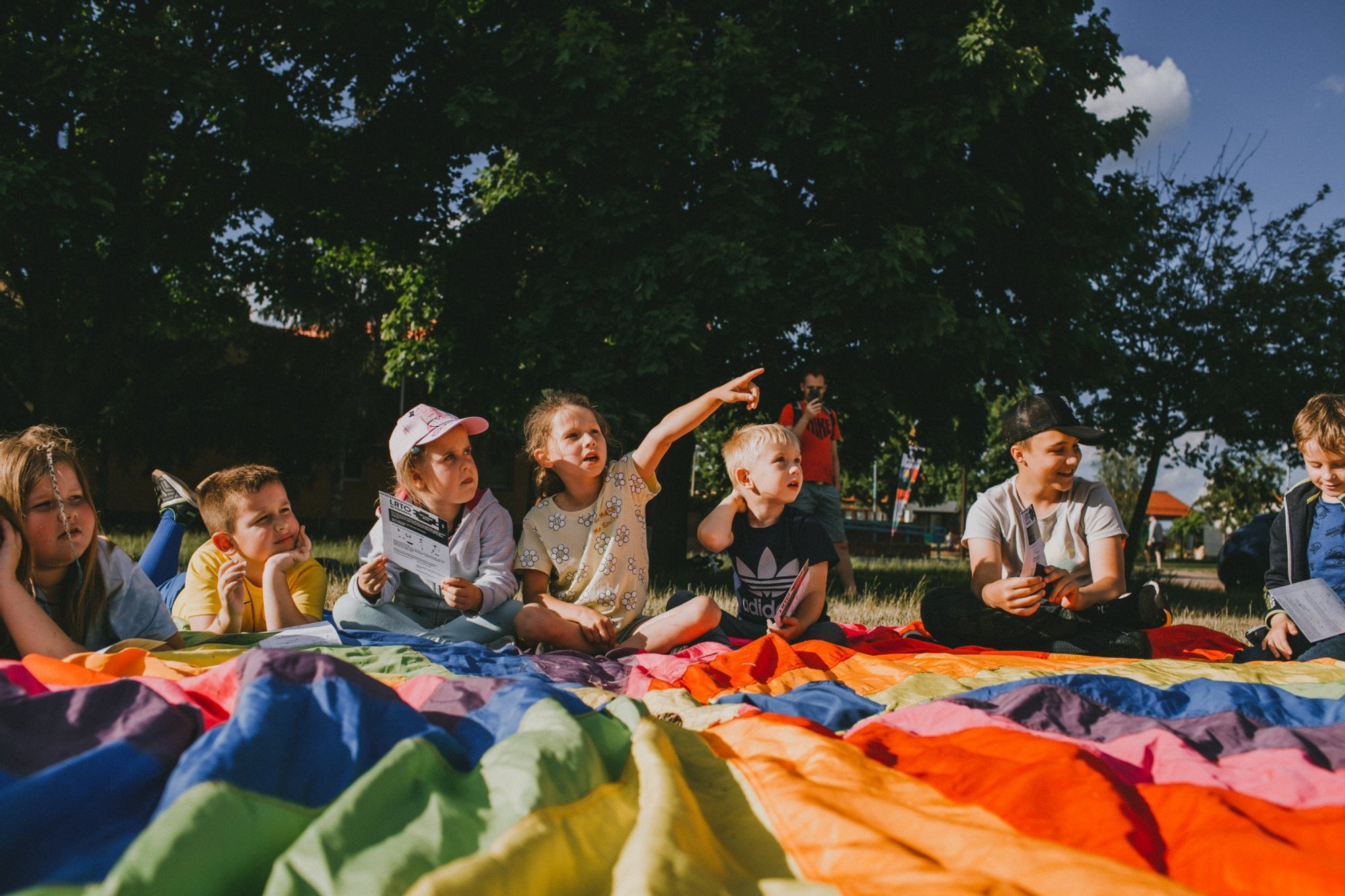 fot. Kalina Żaczek Grupa dzieci w różnym wieku siedzi pod drzewem na kolorowej chuście. Dziewczynka siedząca pośrodku wskazuje palcem poza kadr, reszta dzieci patrzy we wskazanym kierunku.
