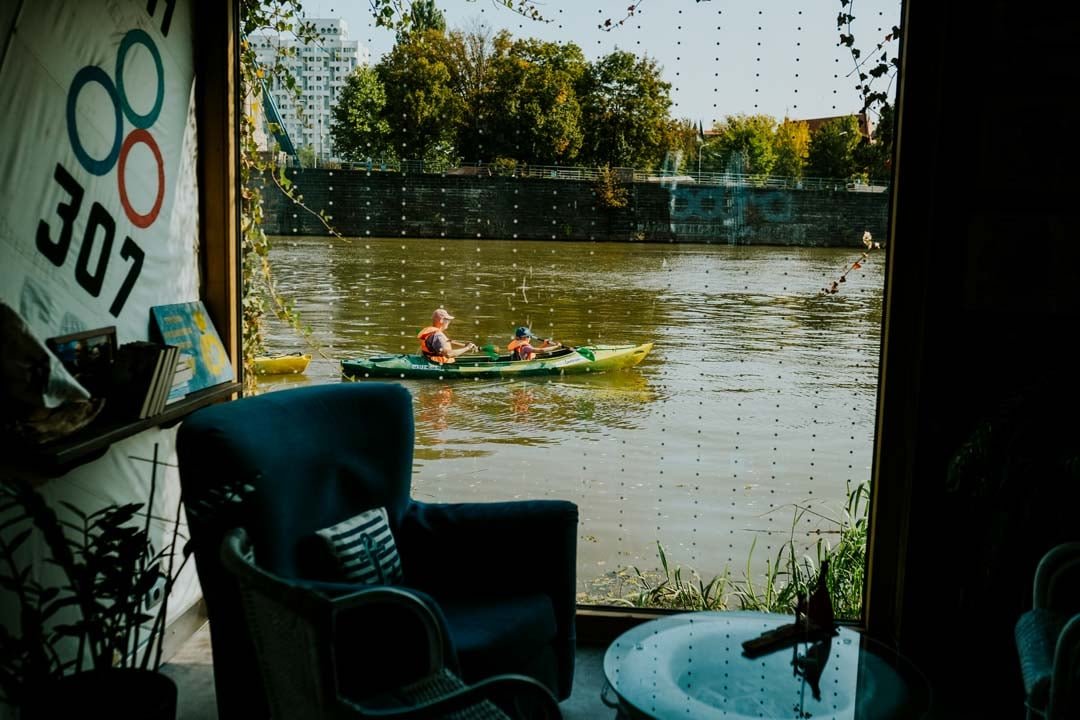 fot. Ewa Jezierska Z wnętrza poieszczenia, w którym stoi duży fotel, widoczna jest rzeka, po której dwie osoby płyną kajakiem.