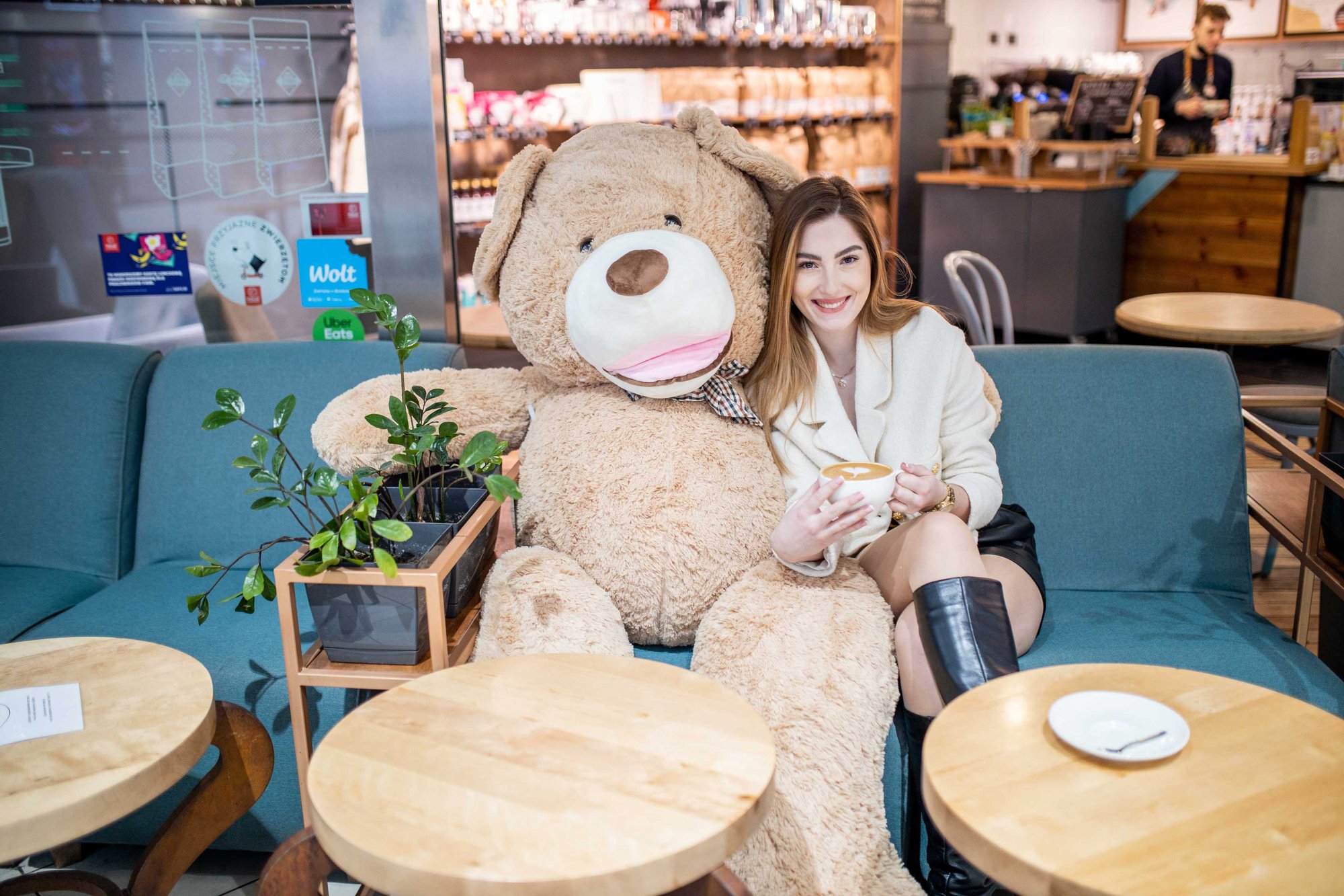 Młoda dziewczyna siedzi na kanapie w kawiarni, w dłoniach trzyma kubek kawy i szeroko się uśmiecha. Obok niej na kanapie siedzi ogromny pluszowy miś. Dziewczyna jest przytulona do misia.