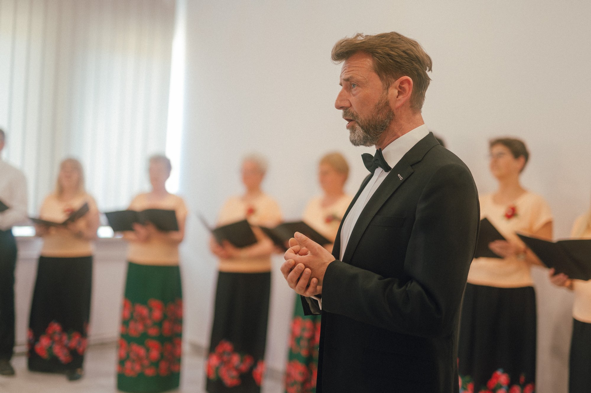 zdjęcie: Marcin Szczygieł Kolorowa fotografia wykonana podczas występu chóru osiedlowego. Zbliżenie na eleganckiego mężczyznę w garniturze - to dyrygent. Za nim stojące w rzędzie chórzystki.