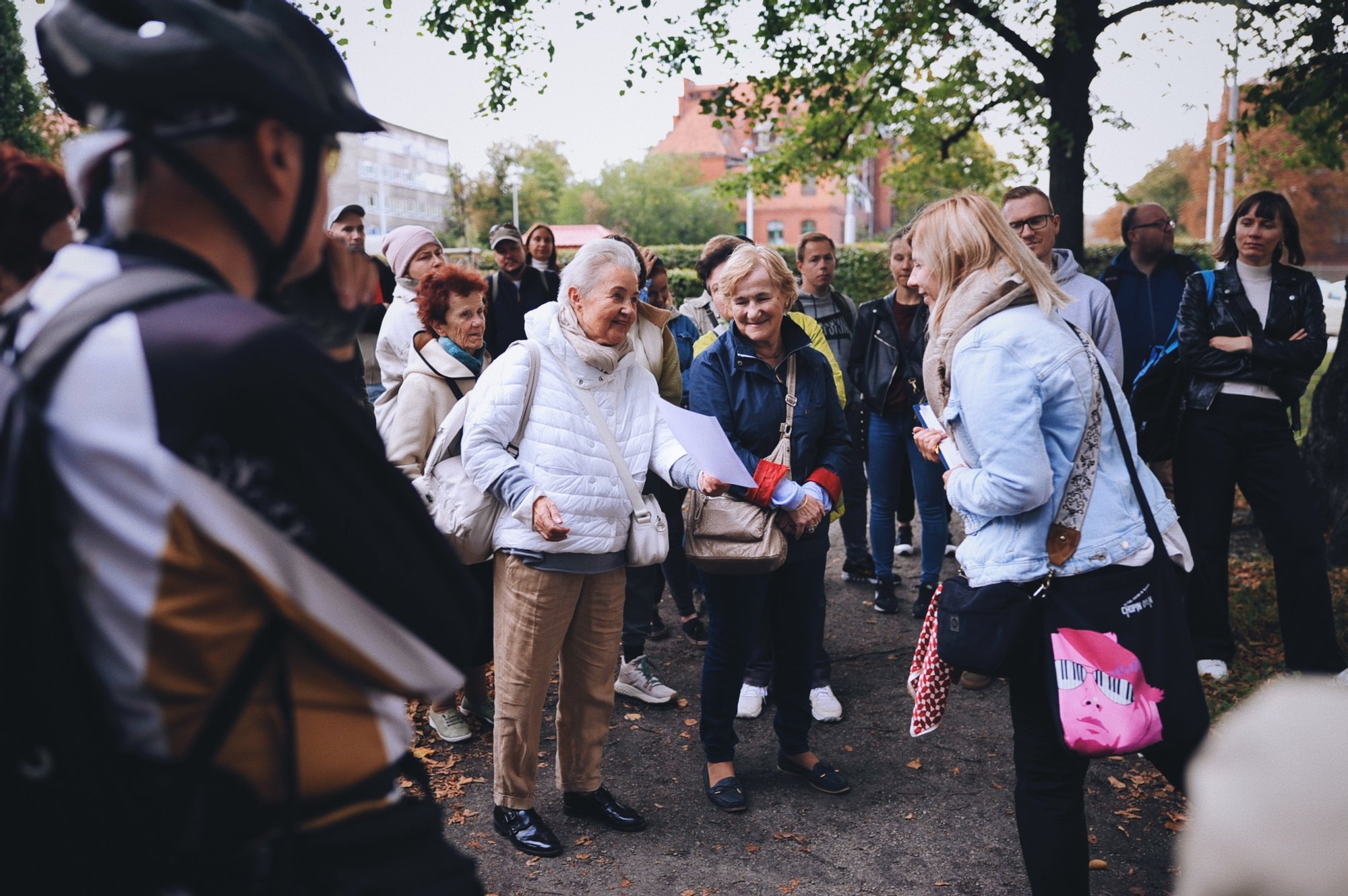 zdjęcie: Anna Pazdej Kolorowa, pozioma fotografia: grupa osób w różnym wieku zebrała się na skwerze. Trwa spacer po osiedlu. Na środku zdjęcia starsza kobieta opowiada historię, pokazując coś na wydrukowanych zdjęciach.