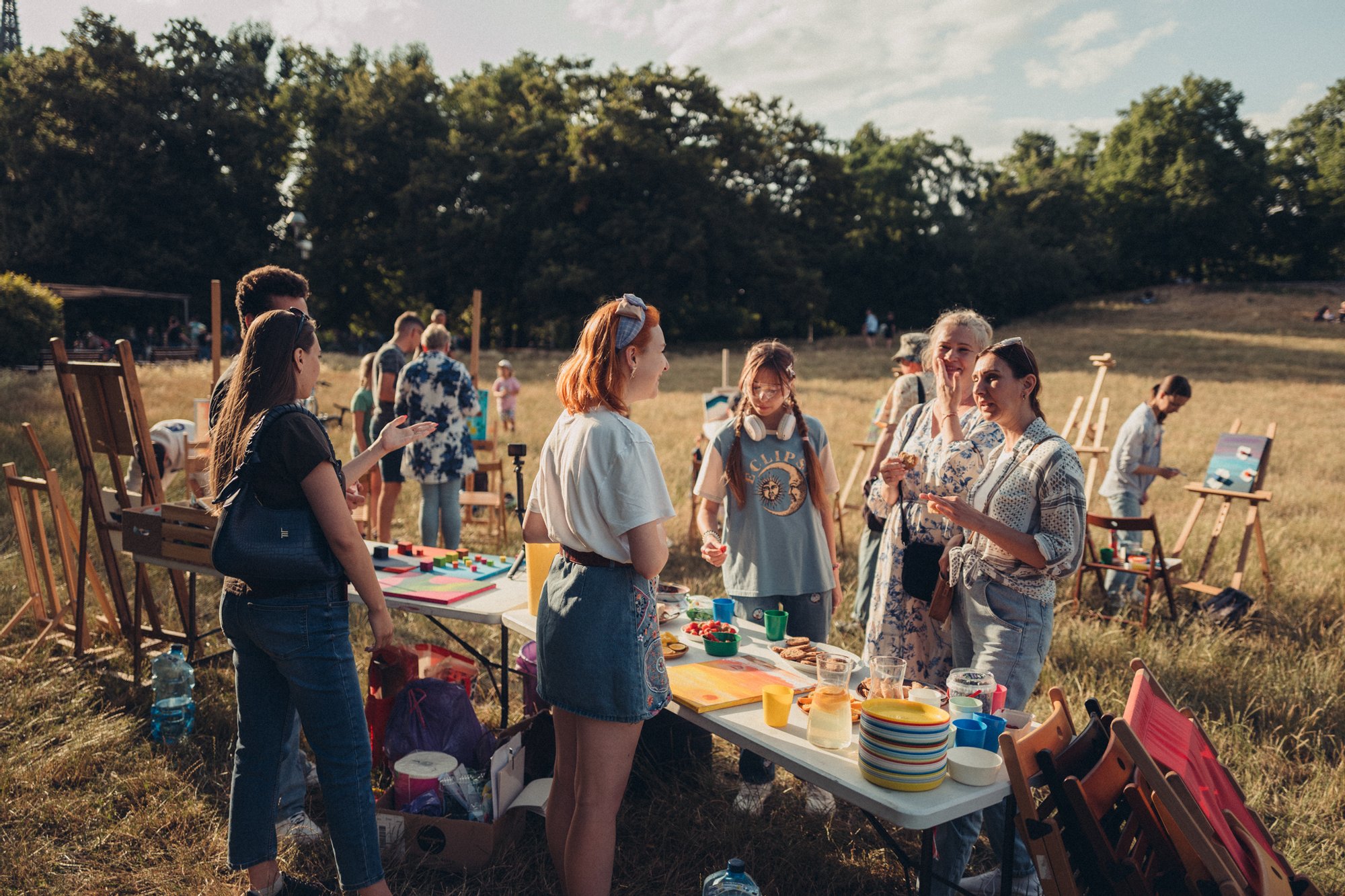 Fot. Wojciech Chrubasik Kolorowa, pozioma fotografia przedstawiająca grupę osób w różnym wieku zebraną na łące w parku. Wszędzie rozstawione są sztalugi i obrazy. W centrum zdjęcia stół z przekąskami/