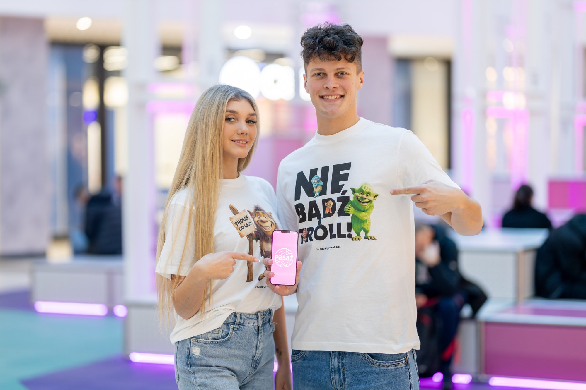 Kolorowa, pozioma fotografia: dwie młode osoby, dziewczyna i chłopak, stoją w Pasażu Grunwaldzkim w koszulkach z napisami "Nie bądź troll" i "Trolle do lasu". Chłopak trzyma w dłoni telefon z logo Pasażu. Oboje wskazują na niego palcami.
