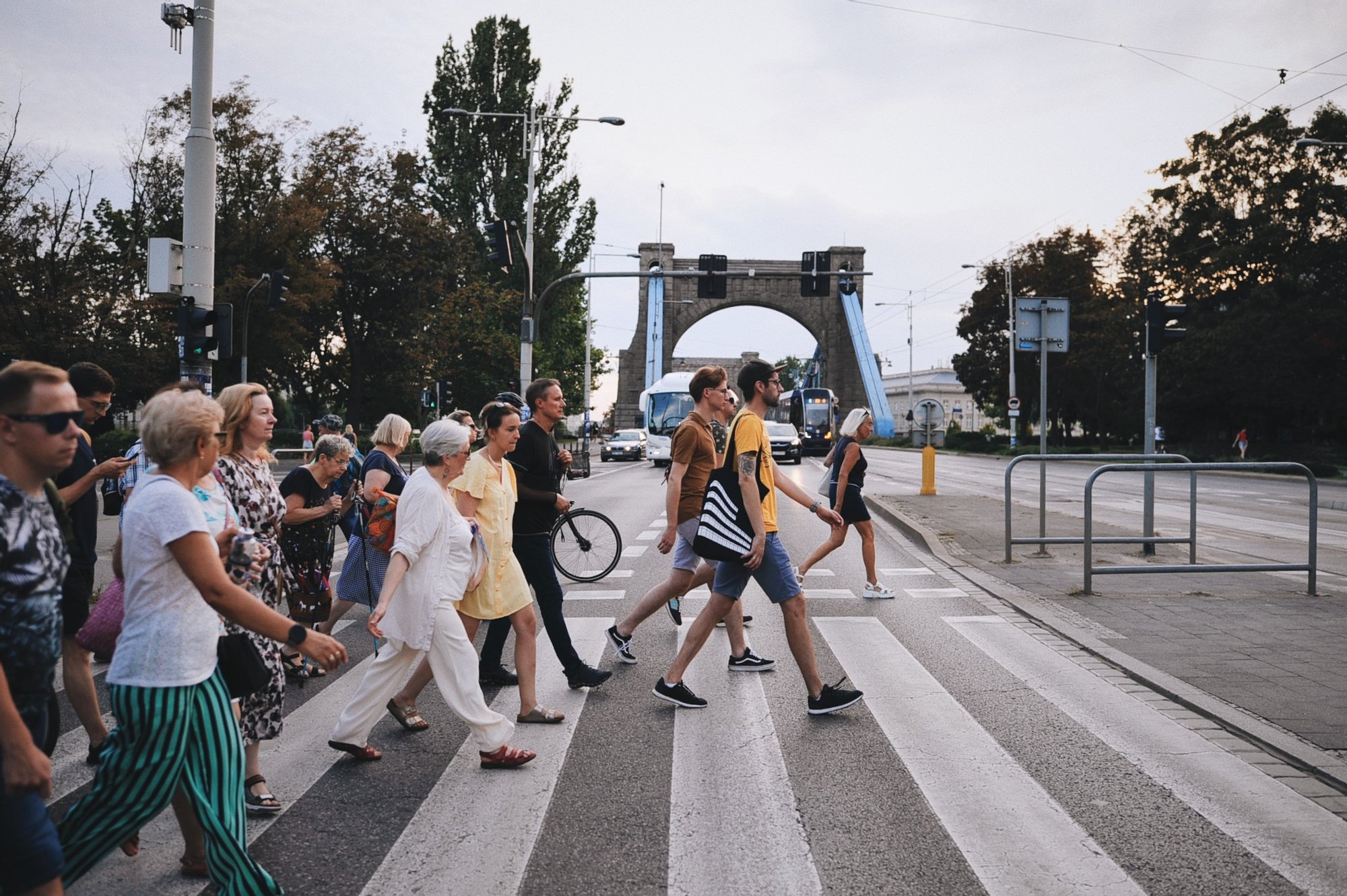 fot. Anna Pazdej Kolorowa fotografia przedstawia dużą grupę osób w różnym wieku przechodzących przez pasy. W tle widoczny jest most Grunwaldzki. Na przedzie grupy idzie Maciek Wlazło - prowadzący spacer po osiedlu.