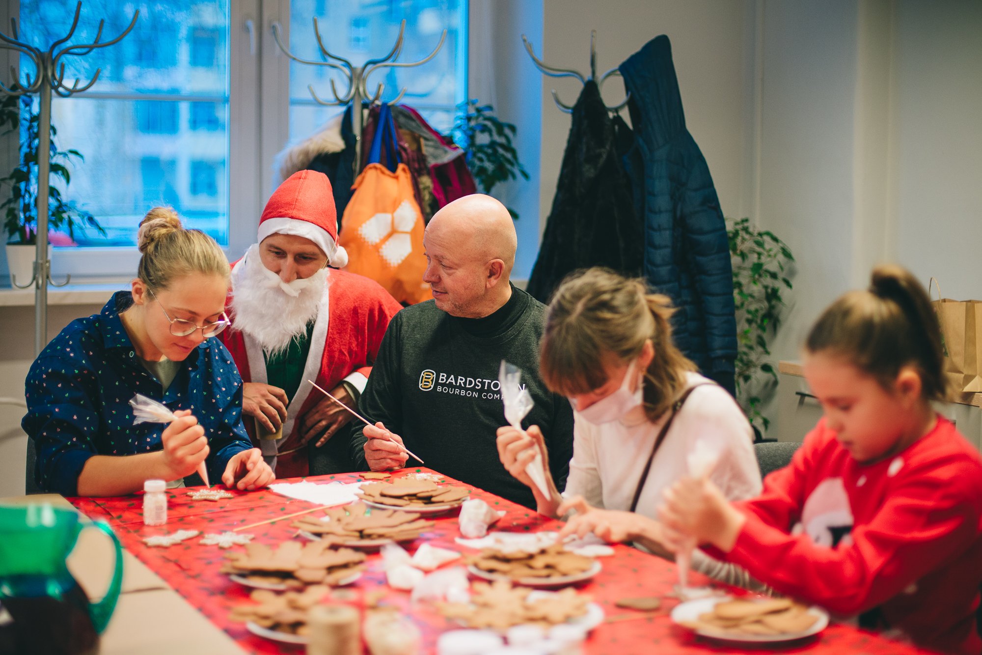fot. Marta Sobala Przy długim stole, przykrytym czerwonym obrusem, stole siedzą osoby w różnym wieku i wspólnie dekorują pierniczki. Razem z nimi siedzi mężczyzna w stroju świętego Mikołaja.