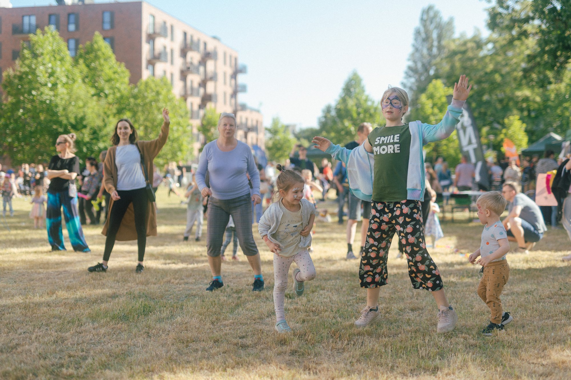 Kolorowa fotografia przedstawia grupę kobiet i dzieci w różnym wieku tańczących na łące. Trwa sąsiedzki piknik.