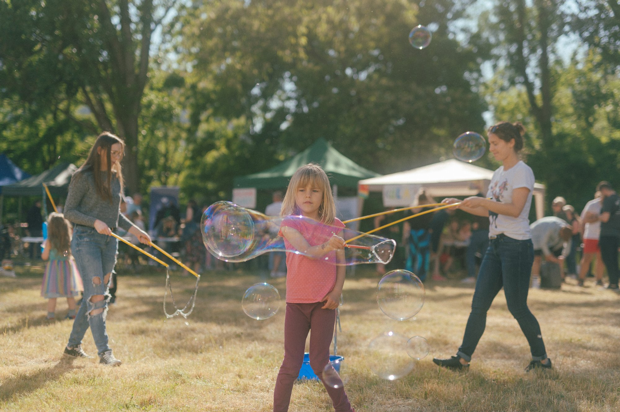 Kolorowa, pozioma fotografia przedstawia kilkuletnią dziewczynkę puszczającą duże bańki mydlane podczas pikniku na łące.