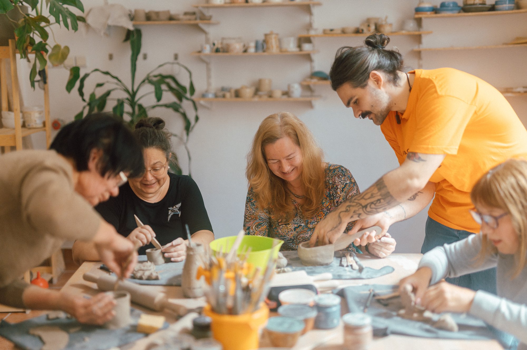 fot. Marcin Szczygieł Kolorowa, pozioma fotografia: pięć seniorek siedzi przy duzym stole i rzeźbi w glinie. Młody mężczyzna pomaga jedne z nich przykleić ucho do kubka.