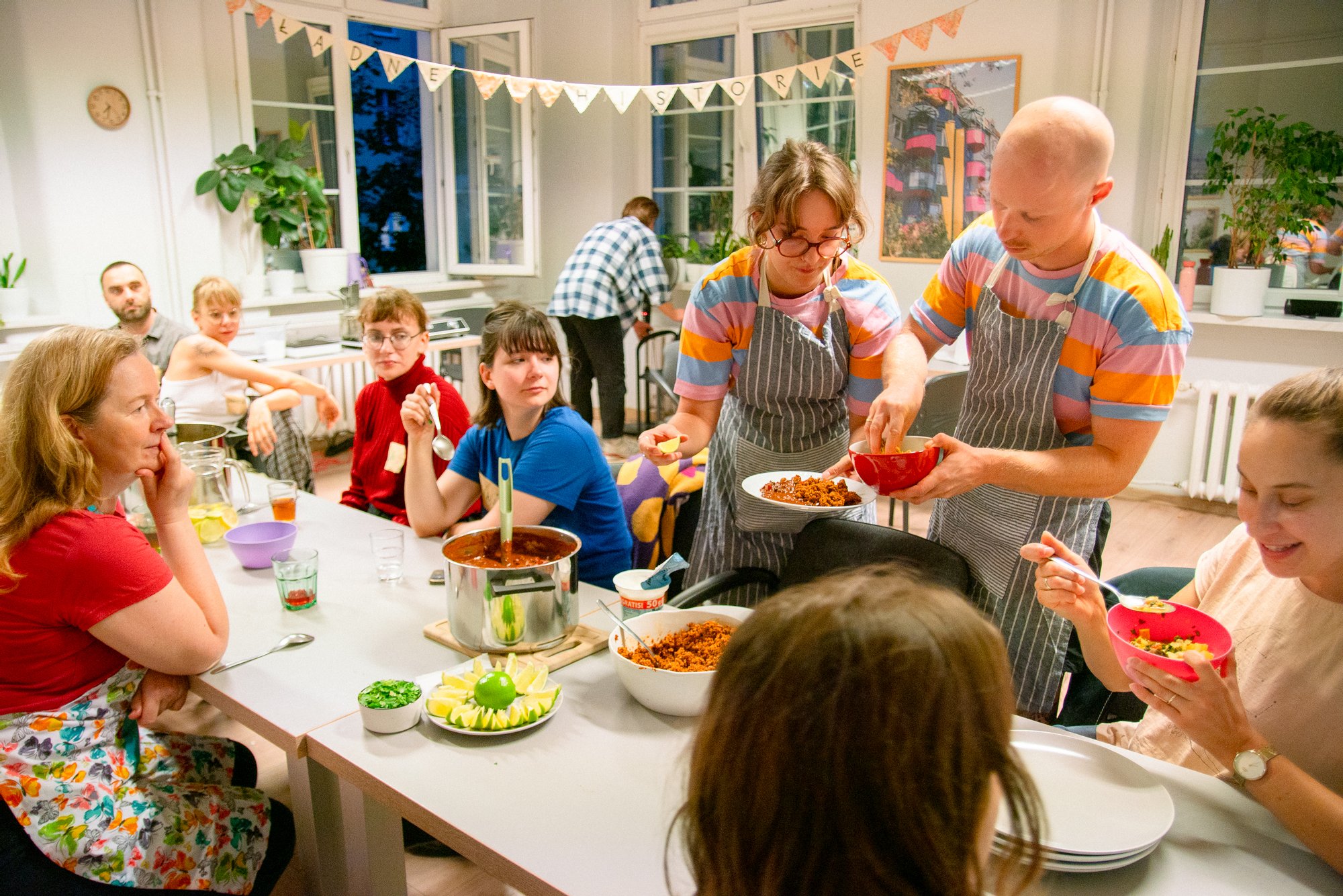 Kolorowa fotografia wykonana podczas warsztatów kulinarnych. W dużym jasnym pomieszczeniu, przy długim stole siedzi grupa osób w różnym wieku. Dwójka młodych ludzi nakłada na talerze przygotowywane wcześniej potrawy.