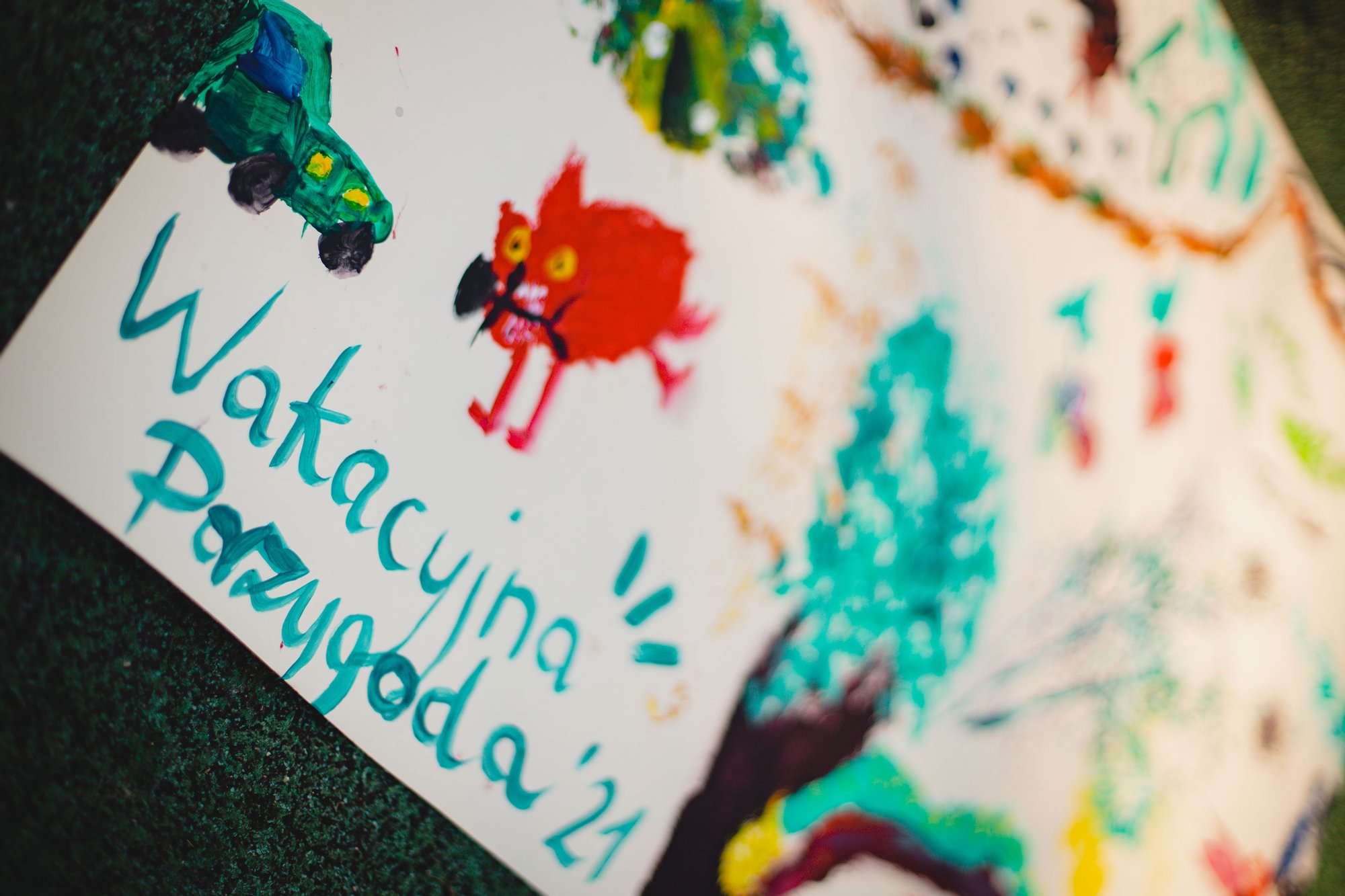 Dziecięcy rysunek przedstawiający zielony samochód, czerwone zwierzątko oraz niebieski napis: Wakacyjna przygoda '21