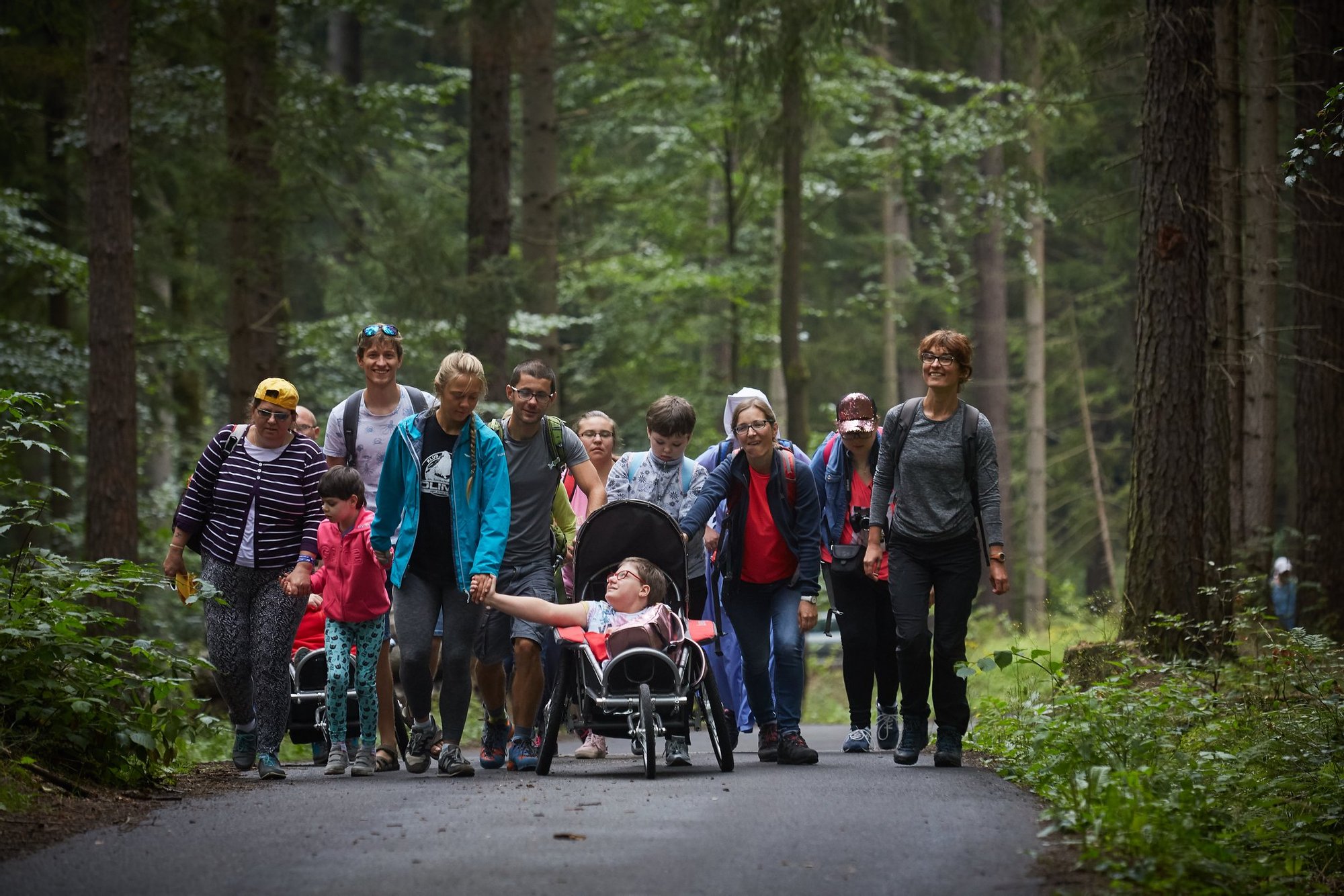 Grupa osób idzie szeroką ścieżką przez las iglasty. Obok siebie idą osoby poruszające się pieszo o własnych siłach oraz w wózkach turystycznych. Pośrodku grupy ide młodza dziewczyna trzymając się za rękę z drugą dziewczyną jadącą w wózku.