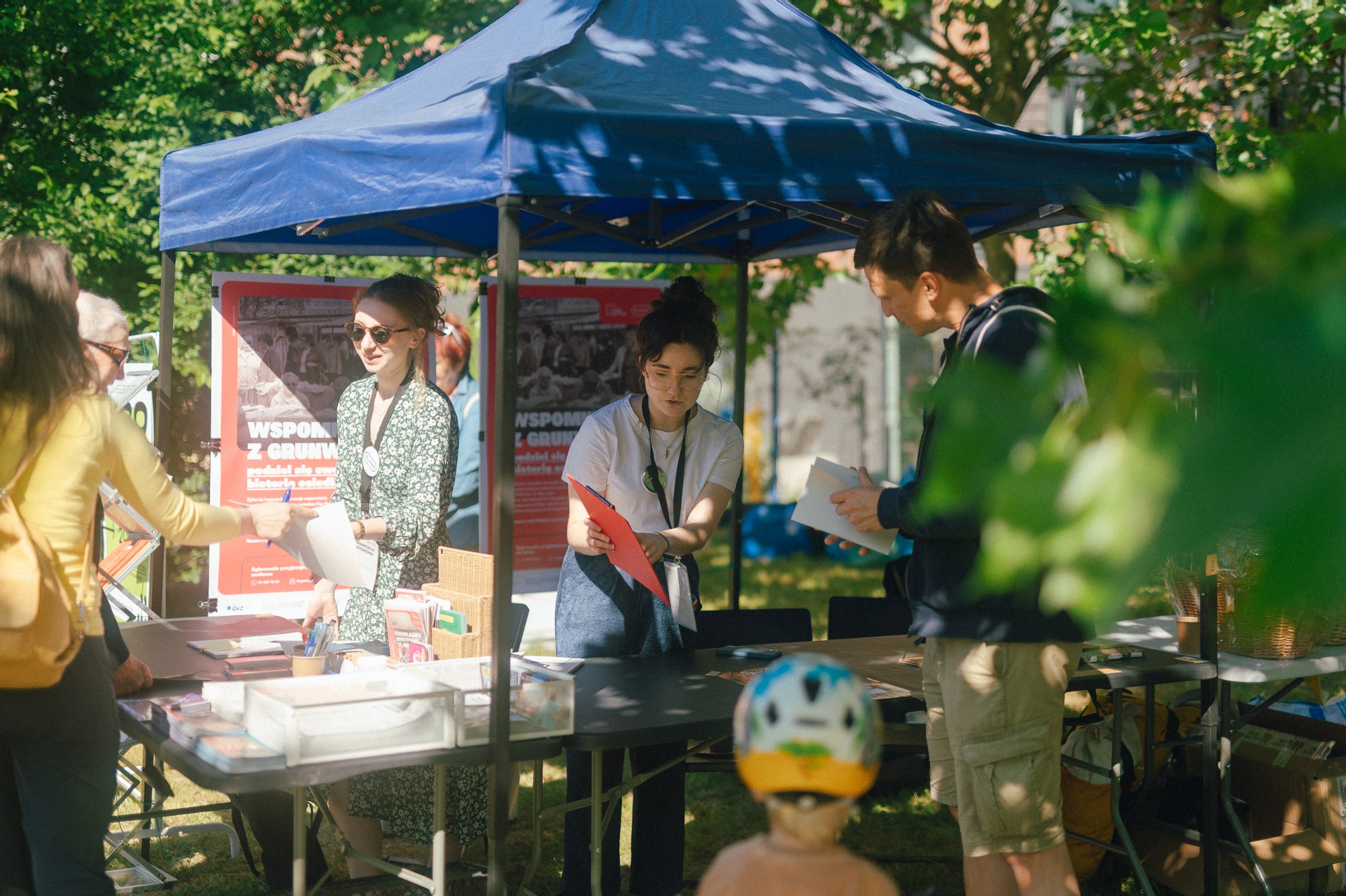 Kolorowa, pozioma fotografia wykonana podczas święta osiedla. Pod namiotem rozstawionym na trawie stoją dwie młode dziewczyny i opowiadają o programie wydarzenia osobom stojącym przy stoisku.
