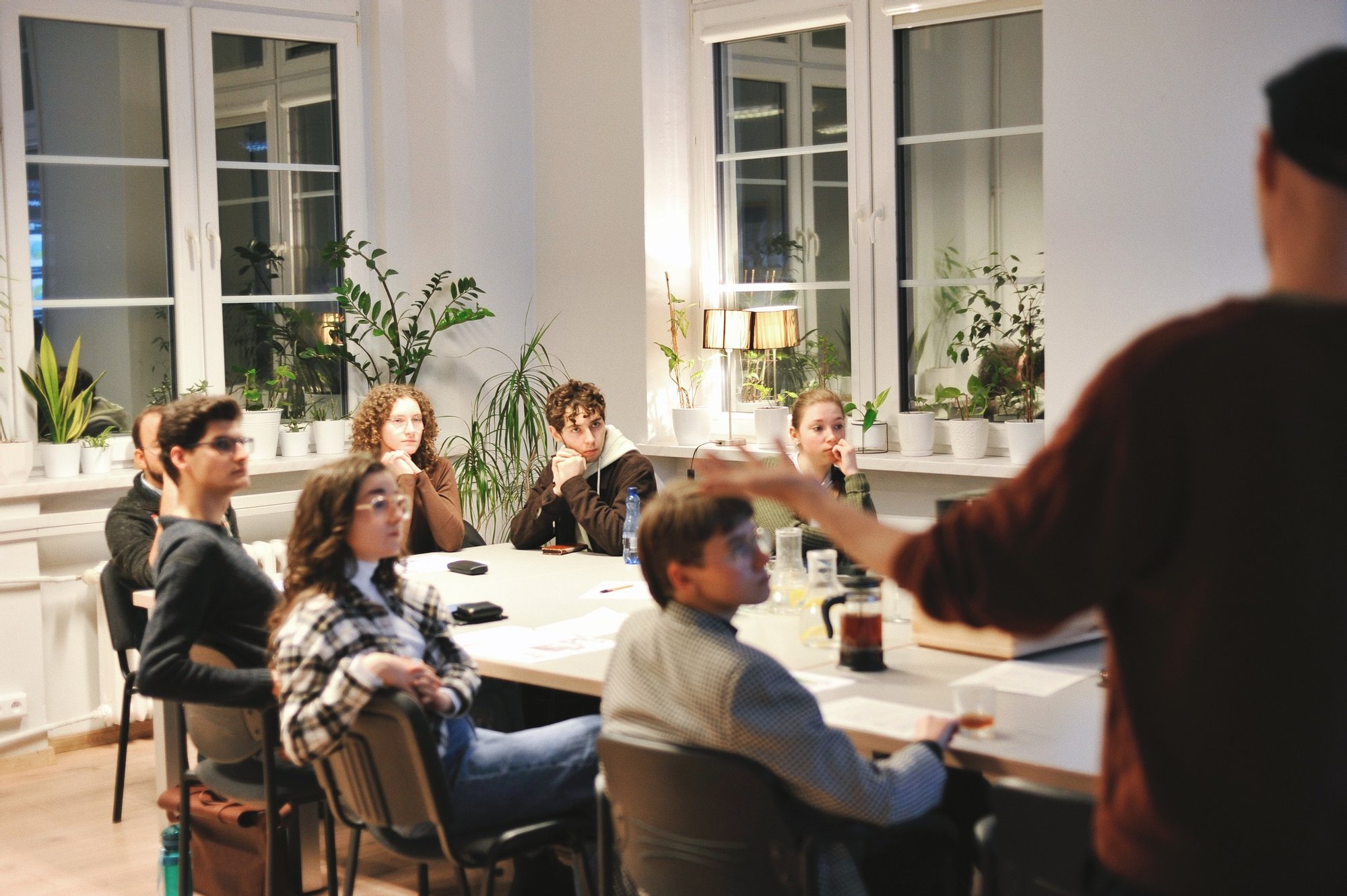 Kolorowa, pozioma fotografia: w jasnym pomieszczeniu przy dużym stole siedzi grupa młodych osób, wszyscy patrzą na ekran znajdujący się poza kadrem. Przy stole stoi mężczyzna i gestykulując opowiada o czymś.