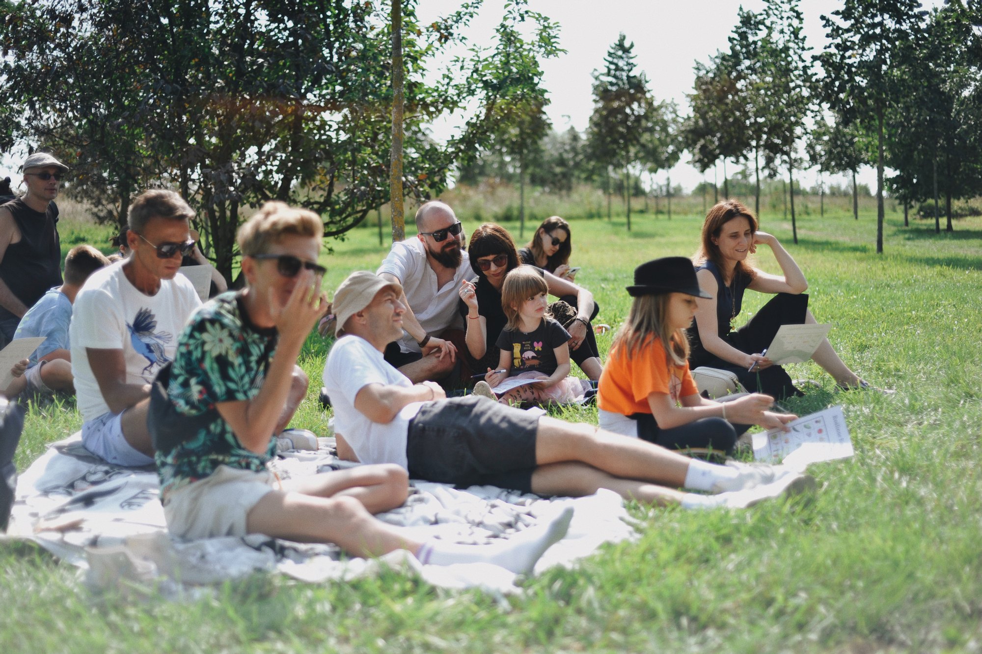 Słoneczny, ciepły dzień. Na zielonej łące porozkładane koce, na których siedzą osoby w różnym wieku. Wokół nich rosną nieduże drzewka.