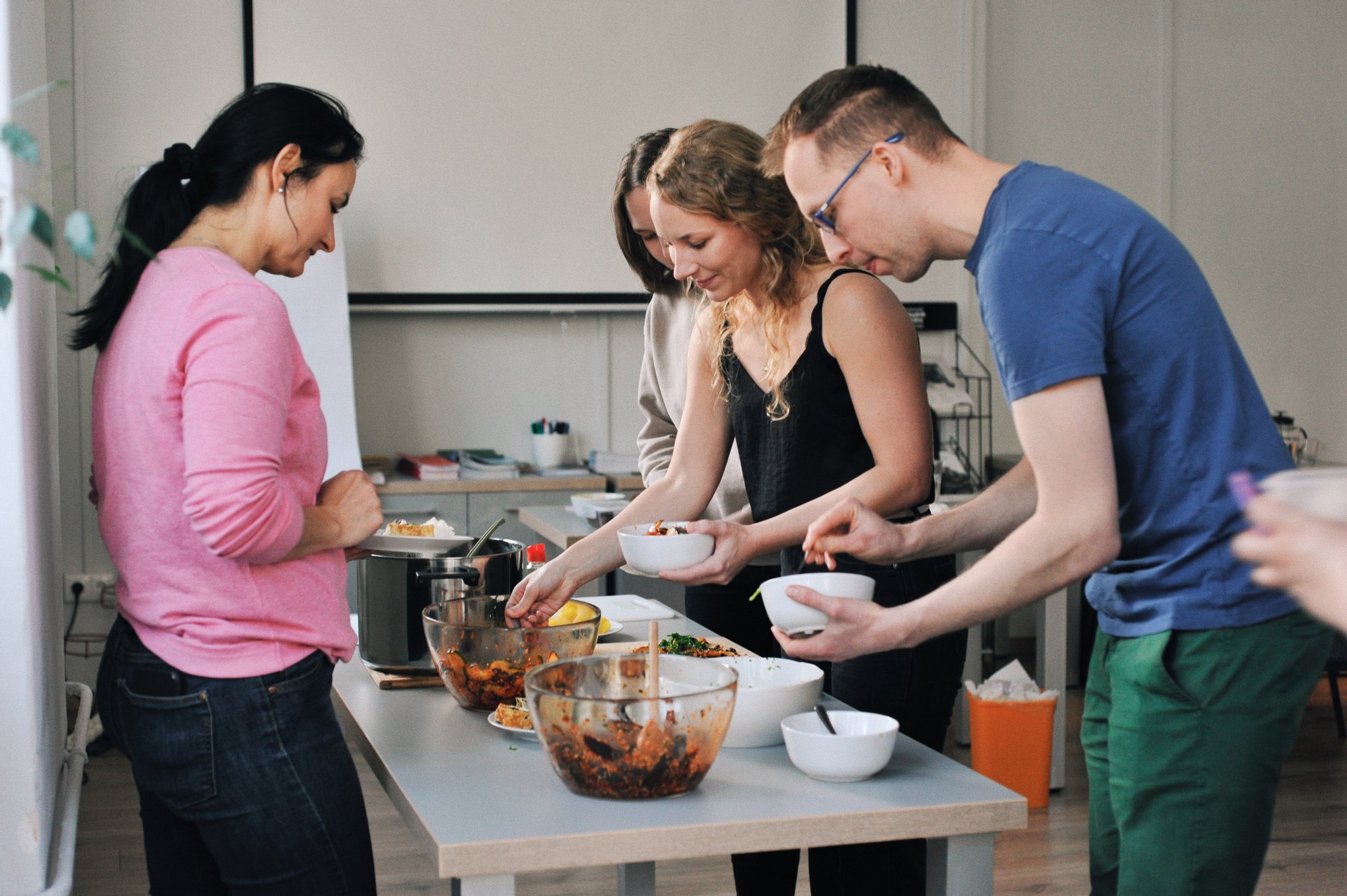 Kolorowa, pozioma fotografia: przy stole warsztatowym w jasnym pomieszczeniu stoją trzy kobiety i mężczyzna. Wspólnie przygotowują posiłek w ramach warsztatów kulinarnych. Na stole stoją miski z półproduktami, naczynia i garnki.