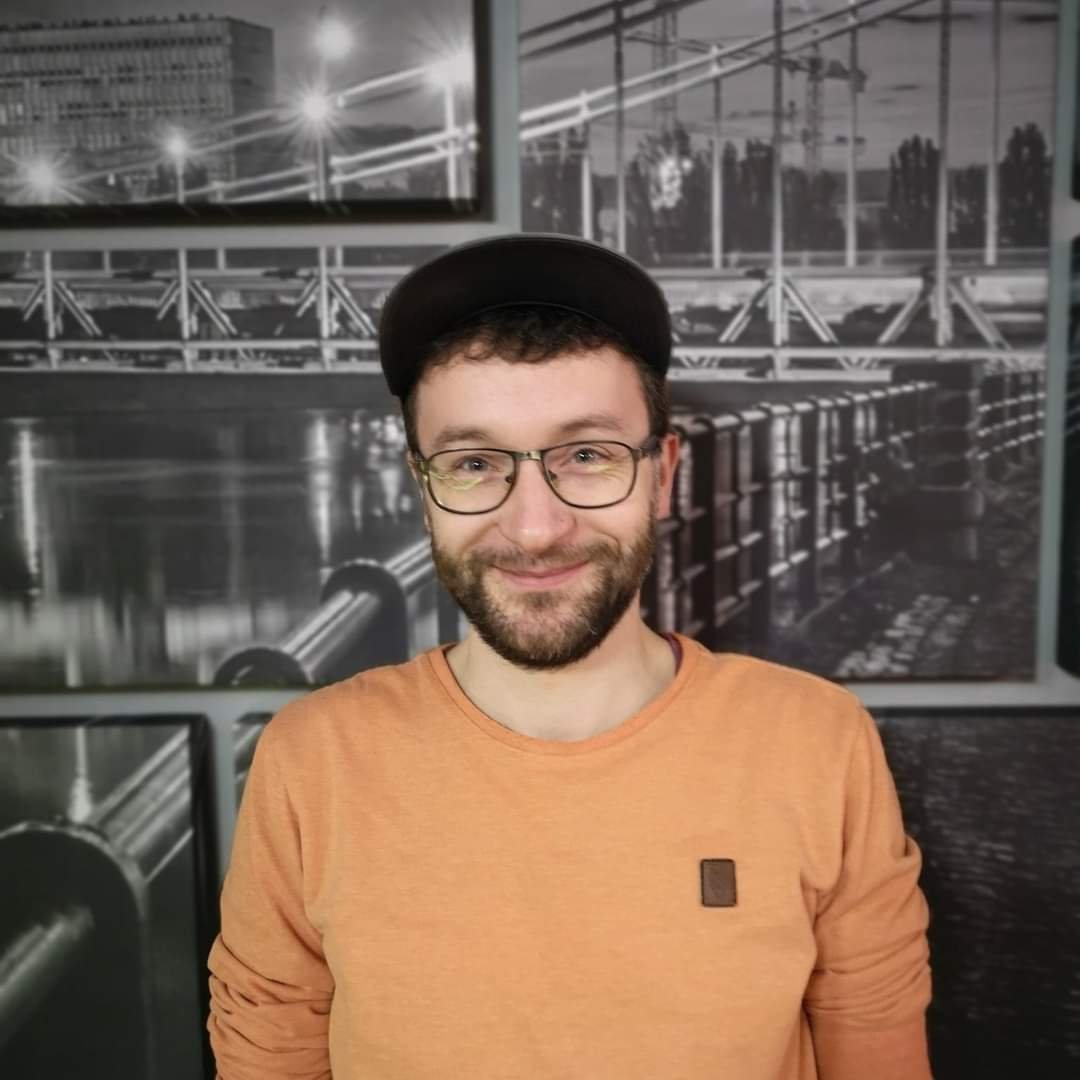 zdjęcie: materiały organizatora Na zdjęciu młody mężczyzna w okularach, czapce z daszkiem i z ciemnym zarostem. Ma pomarańczową koszulkę. Stoi na tle fototapety przedstawiającej Most Grunwaldzki.