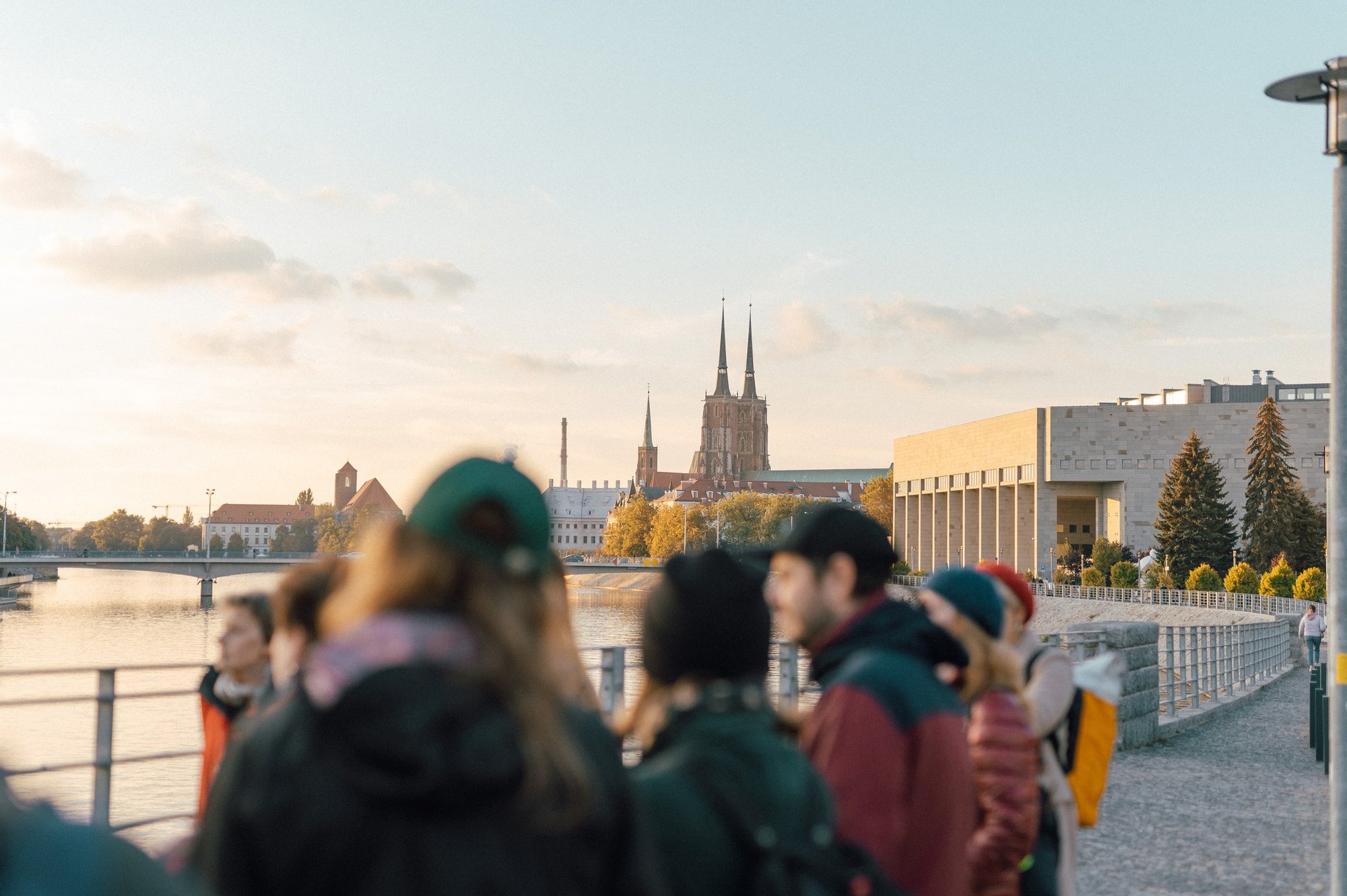 Grupa osób słucha przewodnika, stojąc nad rzeką. Słońce malowniczo zachodz. W tle Biblioteka Uniwersytecka oraz Katedra św. Jana Chrzciciela