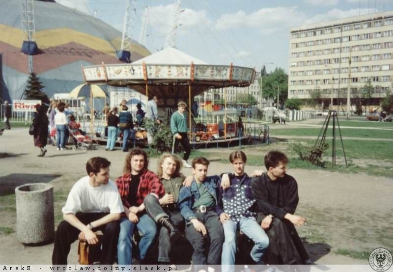 fot. za Wratislaviae Amici Archiwalne, kolorowa fotografia przedstawia grupę nastolatków siedzących przed namiotem cyrkowym na ławce. W tle widać kolorową karuzelę.