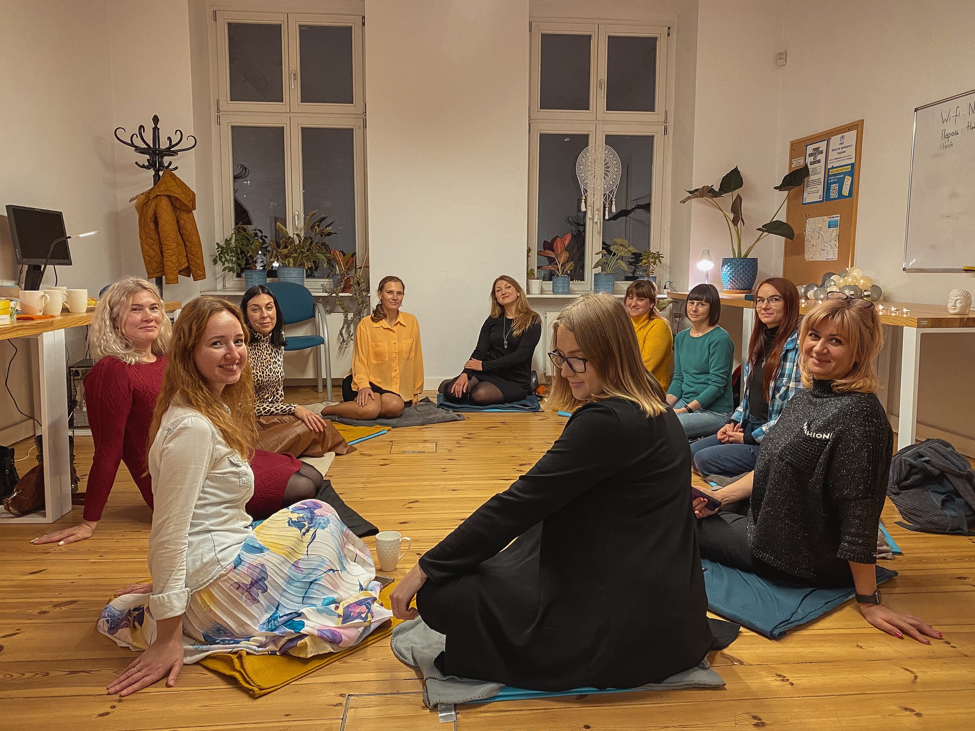fot. Tatiana Kit Na podłodze w kręgu siedzi grupa kobiet w różnym wieku. Spoglądają w obiektyw i uśmiechają się. To uczestniczki kręgu kobiet.