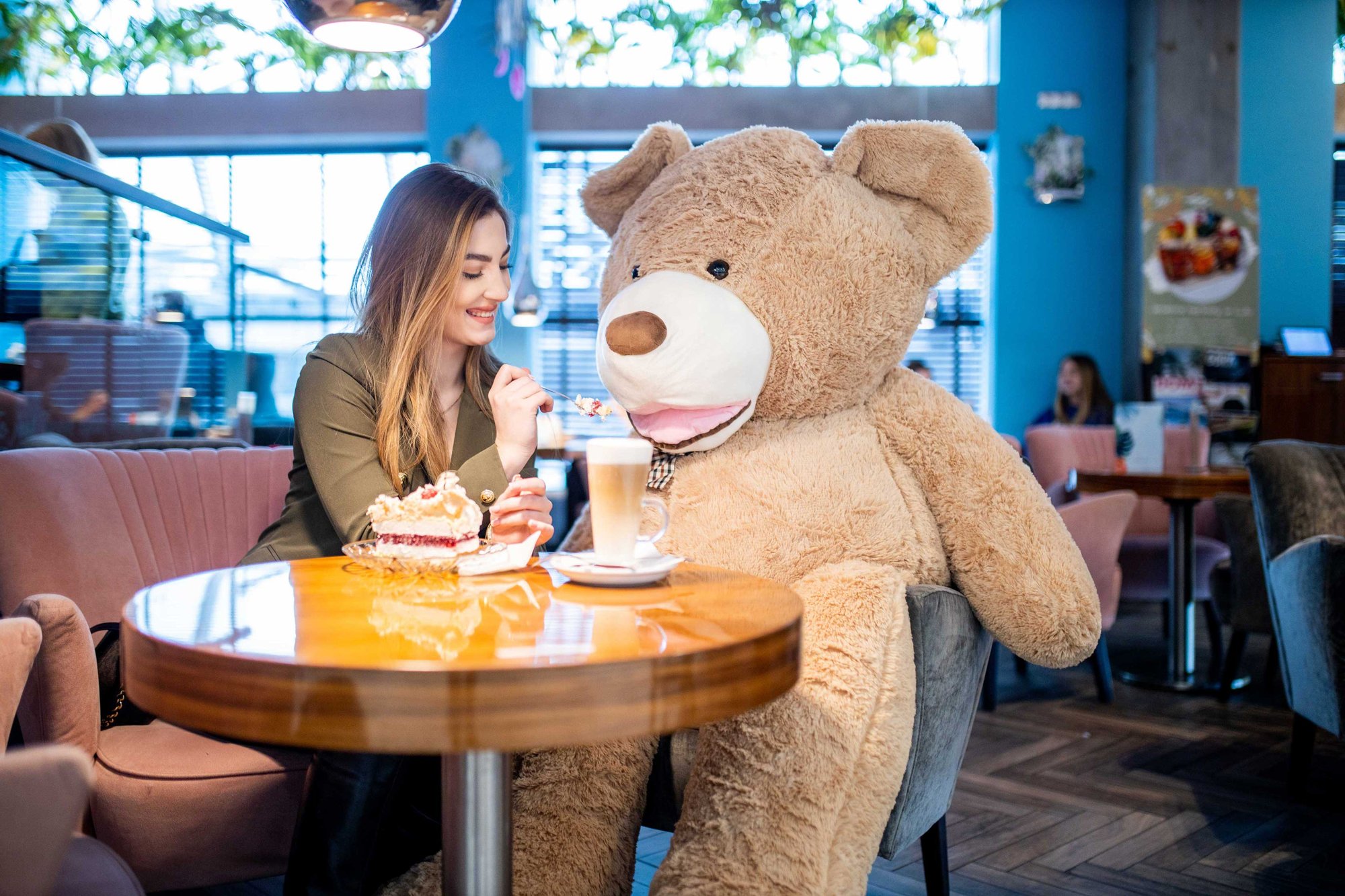 Młoda dziewczyna siedzi w kawiarni, przed nią na stoliku stoi kawa oraz duży kawałek tortu. Na fotelu obok dziewczyny siedzi wielki pluszowy miś. Dziewczyna karmi misia tortem.