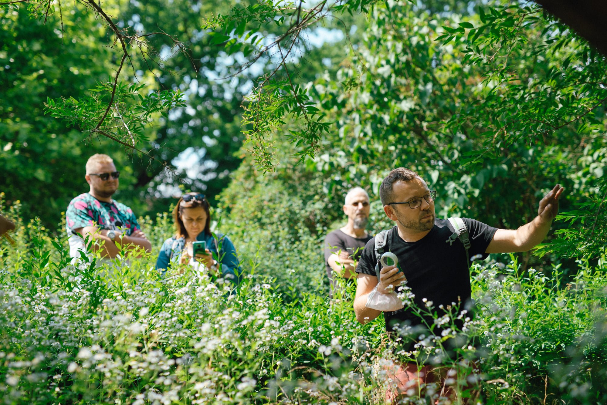 fot. Marcin Szczygieł Spacer z Robertem Sobolewskim - dwóch mężczyzn i kobieta stoją pośród wysokich traw i krzewów za przewodnikiem, który wskazuje coś ręką i opowiada. W drugiej dłoni trzyma lupę.