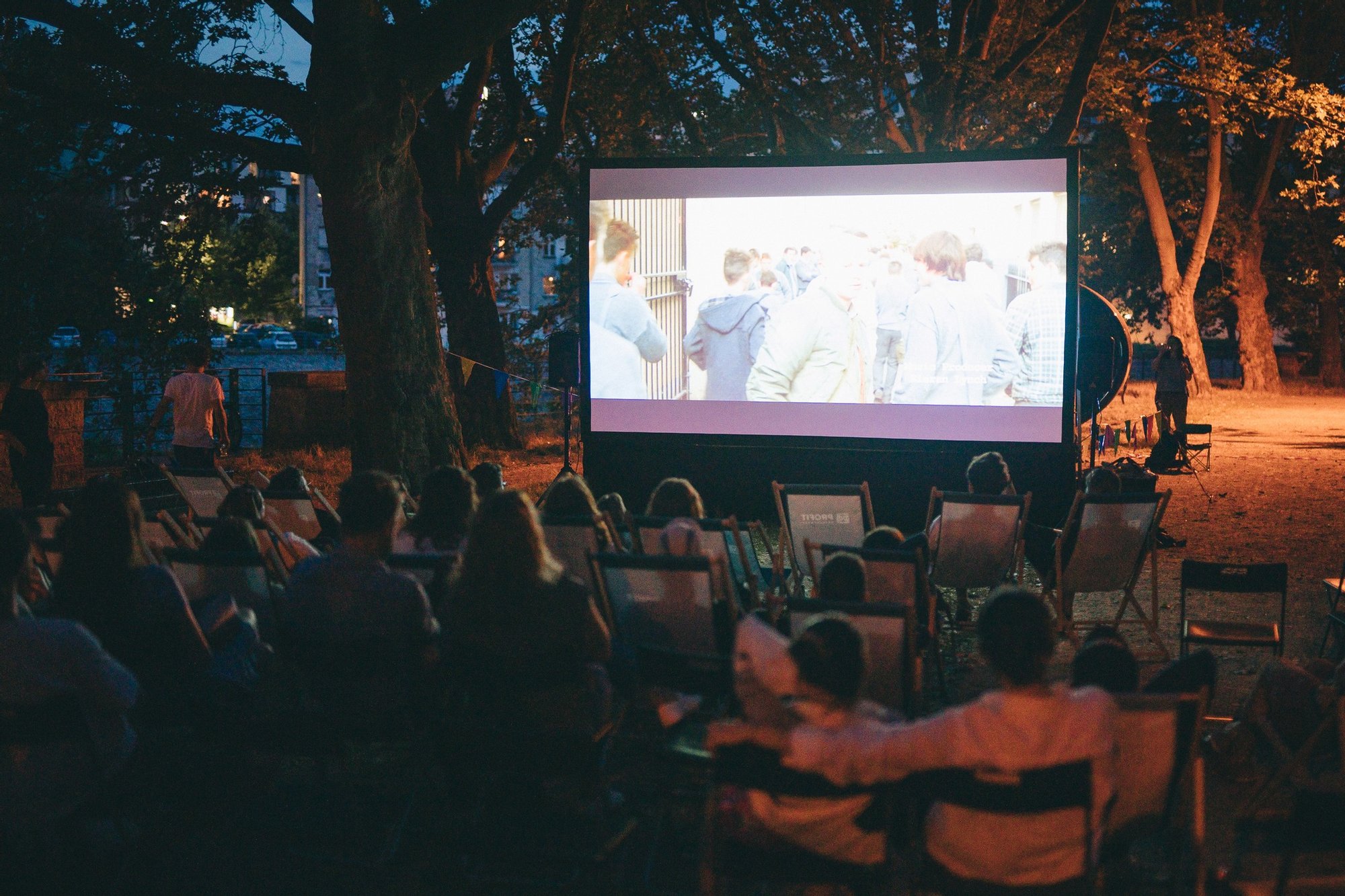 Kolorowa fotografia: duża grupa osób w różnym wieku siedzi na leżakach pod drzewem i ogląda plenerowy pokaz filmu.