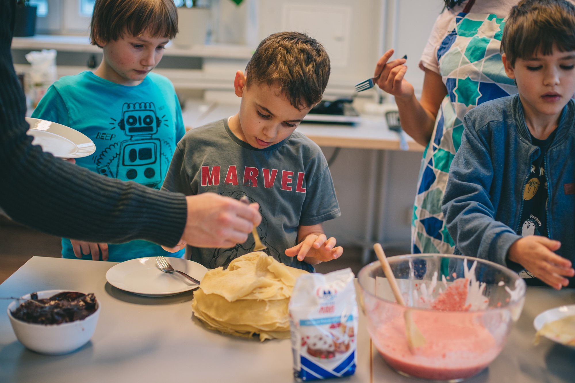 Фотографії: Marta Sobala Trójka kilkuletnich chłopców stoi przy stole i dekorują naleśniki. Na stole stoi talerz z naleśnikami, miska z kremem czekoladowym oraz duża miska z różową polewą.