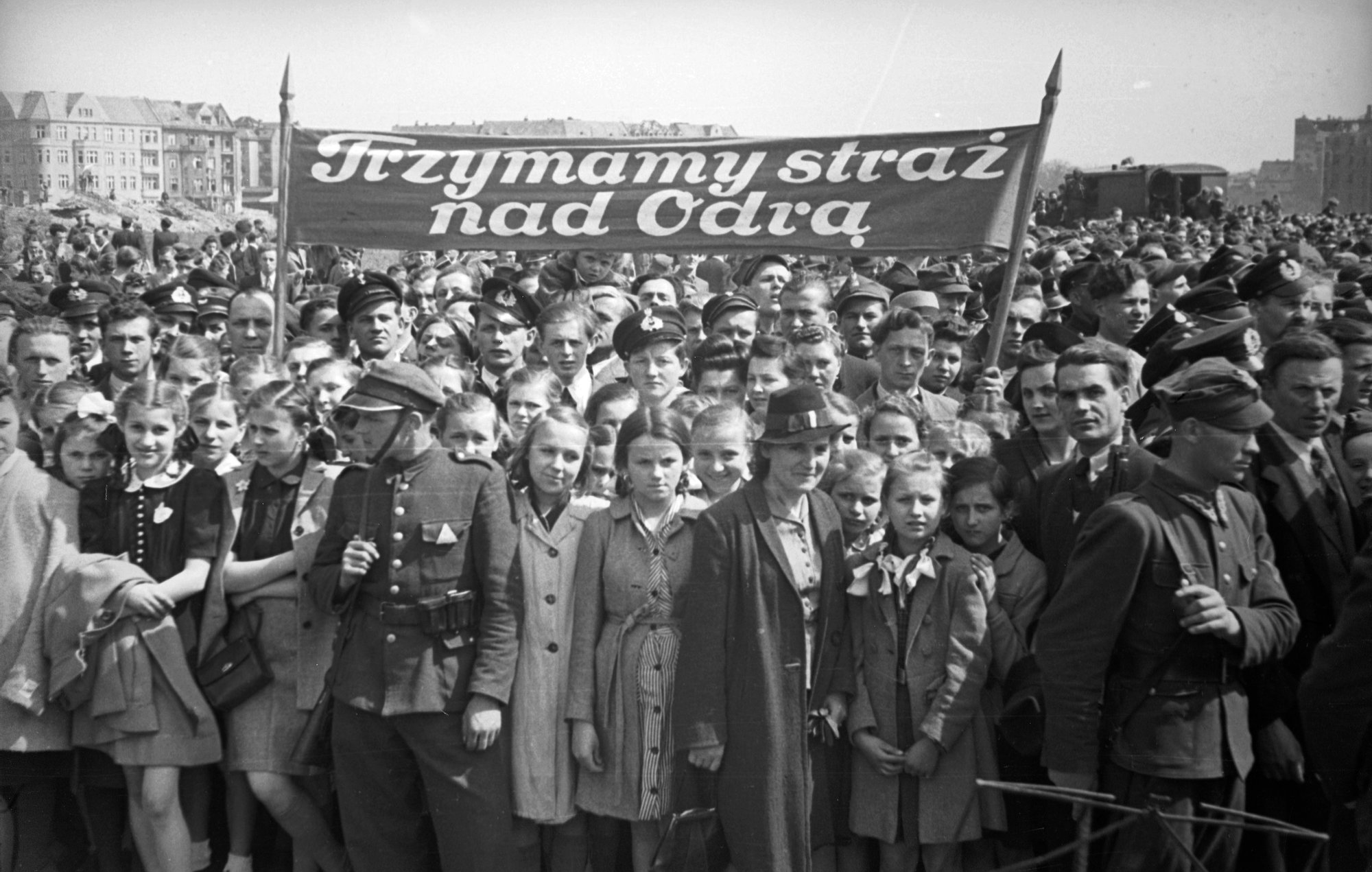 Archiwalne, czarno-białe zdjęcie. Manifestacja na placu Grunwaldzkim w latach czterdziestych XX wieku – w tle kamienice przy ul. Grunwaldzkiej. Na pierwszym planie młodzież trzymająca transparent z napisem: trzymamy straż nad Odrą. Przed nimi stoją żółnierze.