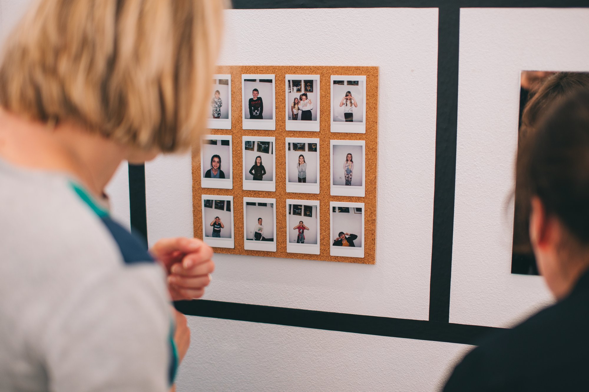 Dwie osoby oglądają zdjęcia na wystawie. Widzimy tylko fragmenty ich sylwetek. Osoba po lewej ma krótkie jasne włosy i jasny sweter, osoba po prawej ma ciemne długie włosy i ciemne ubranie.