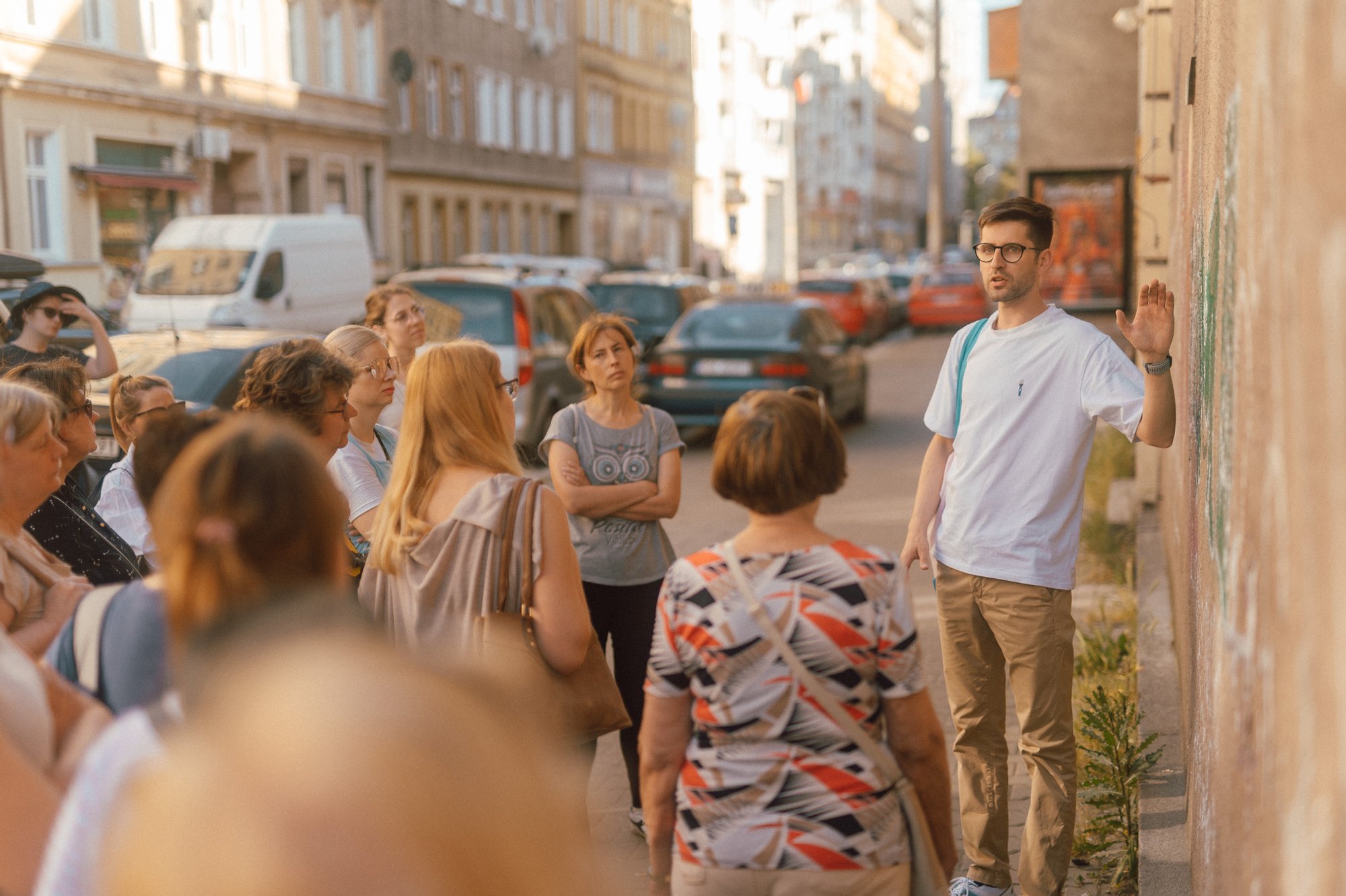 zdjęcie: Marcin Szczygieł Kolorowa, pozioma fotografia, przedstawiająca grupę osób w różnym wieku, stojącą na chodniku. Przed nimi Maciek Wlazło - przewodnik spaceru, opowiadający o miejscu, w którym przystanęła grupa.